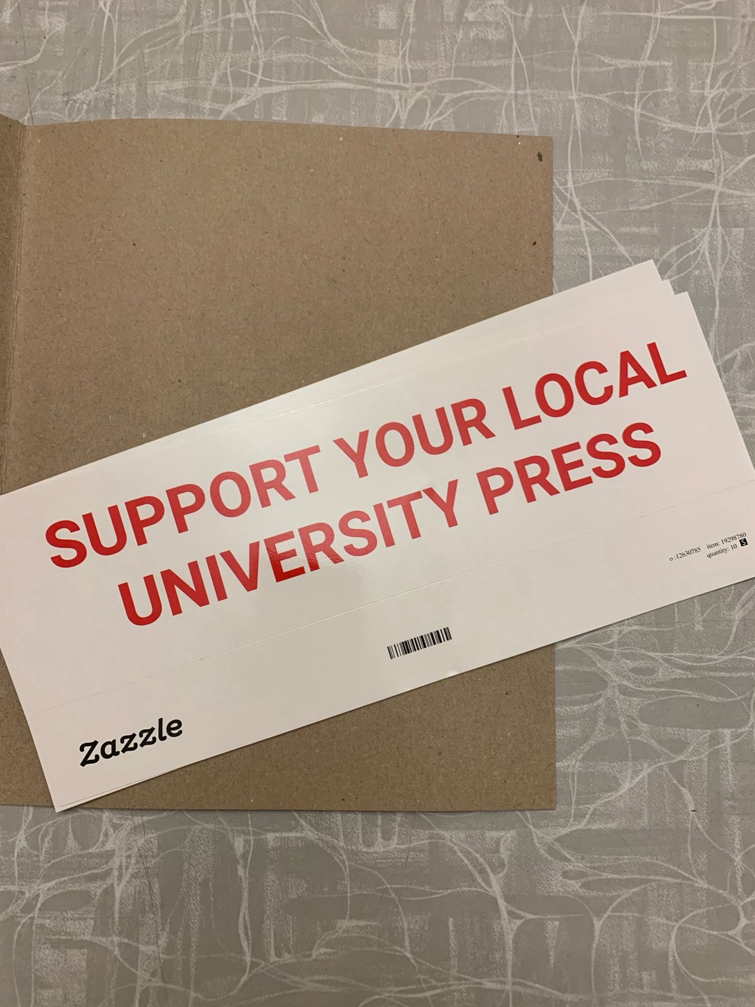 White bumper sticker, sitting on a table. Sticker reads “SUPPORT YOUR LOCAL UNIVERSITY PRESS.”