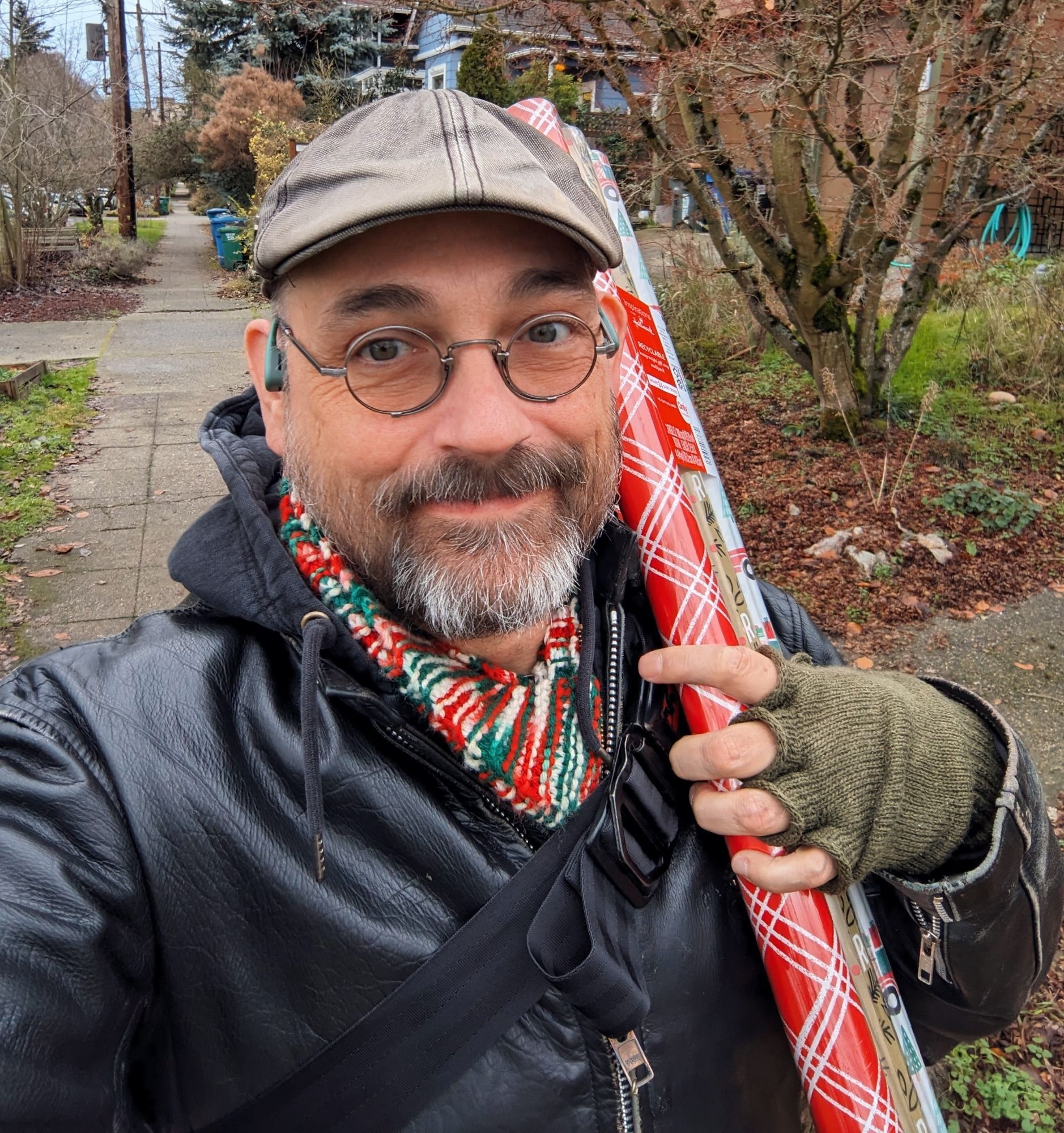 Selfie of me, a white cisgay dude, holding wrapping paper tubes and wearing a crocheted scarf with Christmas colors (red, green, white).