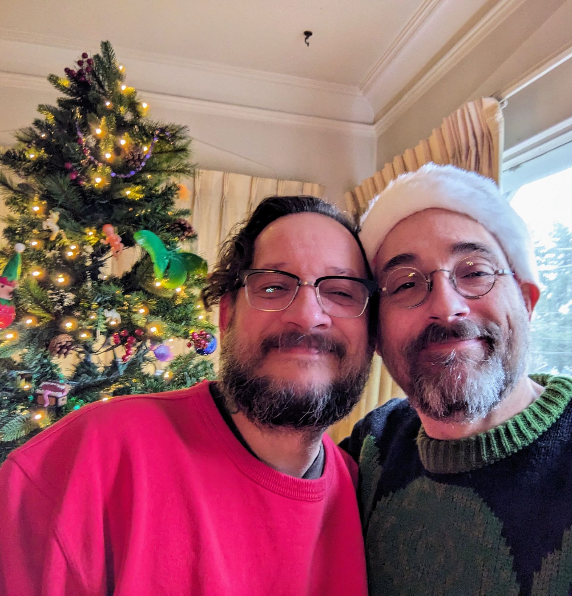 Two scruffy cisgay white dudes mugging for the camera while standing in front of a Christmas tree.