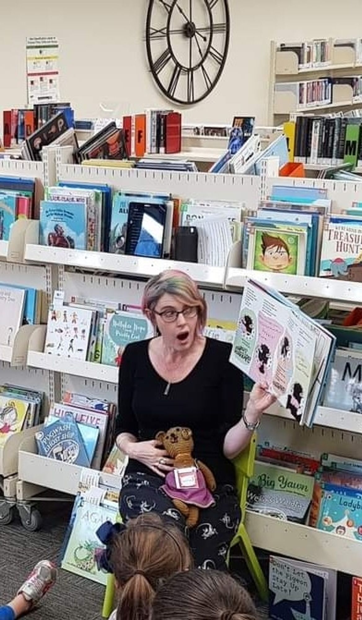 A Librarian, in black pyjamas with dinosaur skeletons, is expressively reading Chocolate Cake by Michael Rosen  
She is holding a handknitted teddy bear, who is wearing a purple skirt. She has one arm.