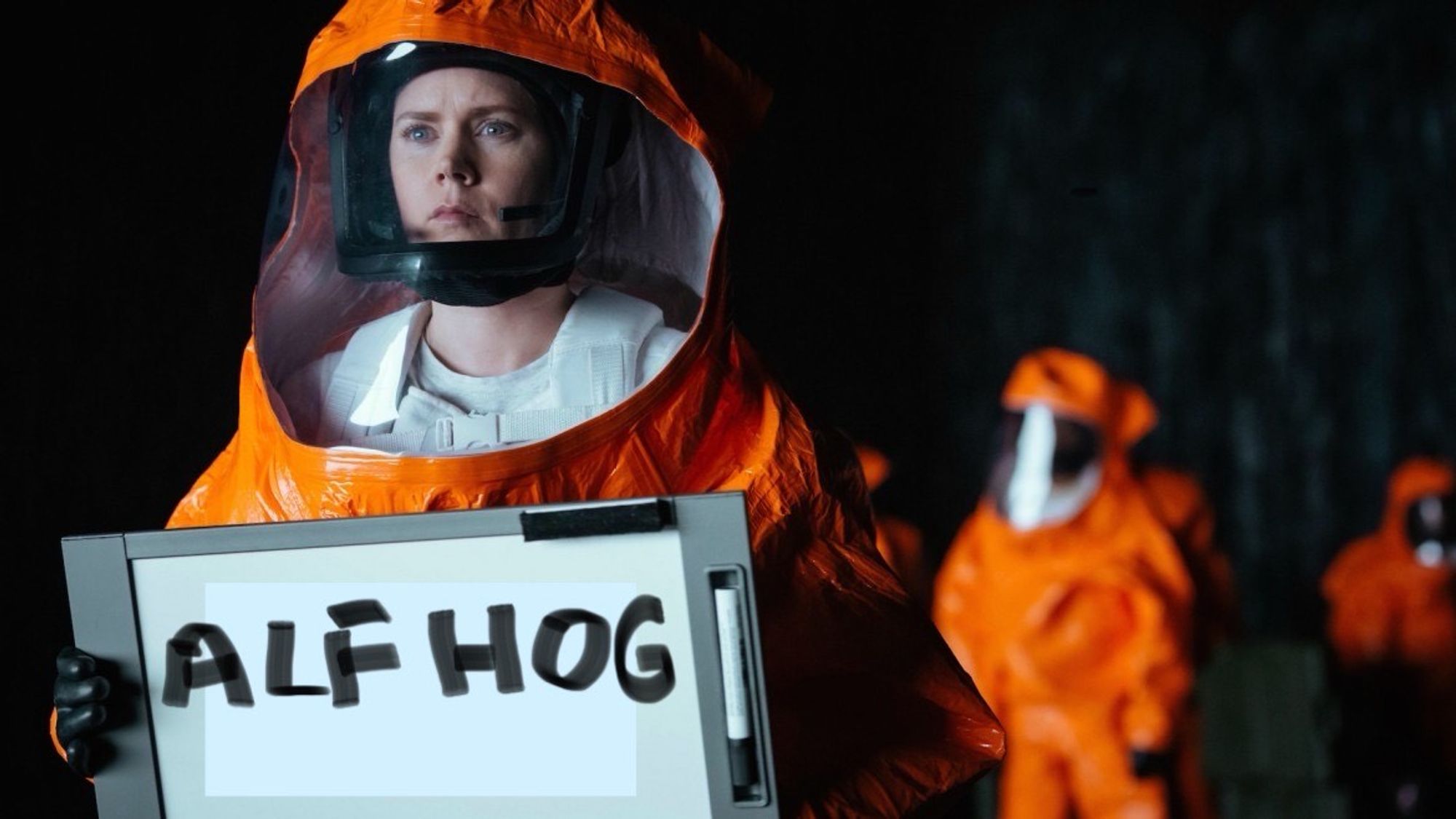 Scene from movie Arrival, woman in orange hazmat suit holding up whiteboard with words “ALF Hog.”