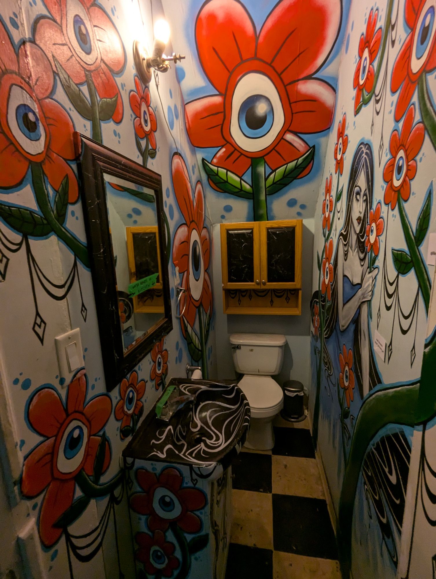 A narrow bathroom decorated with red flowers centered around a blue irised eye. A white skinned woman with long black hair, white eyes in a strapless blue dress holds one smaller flower to her face.