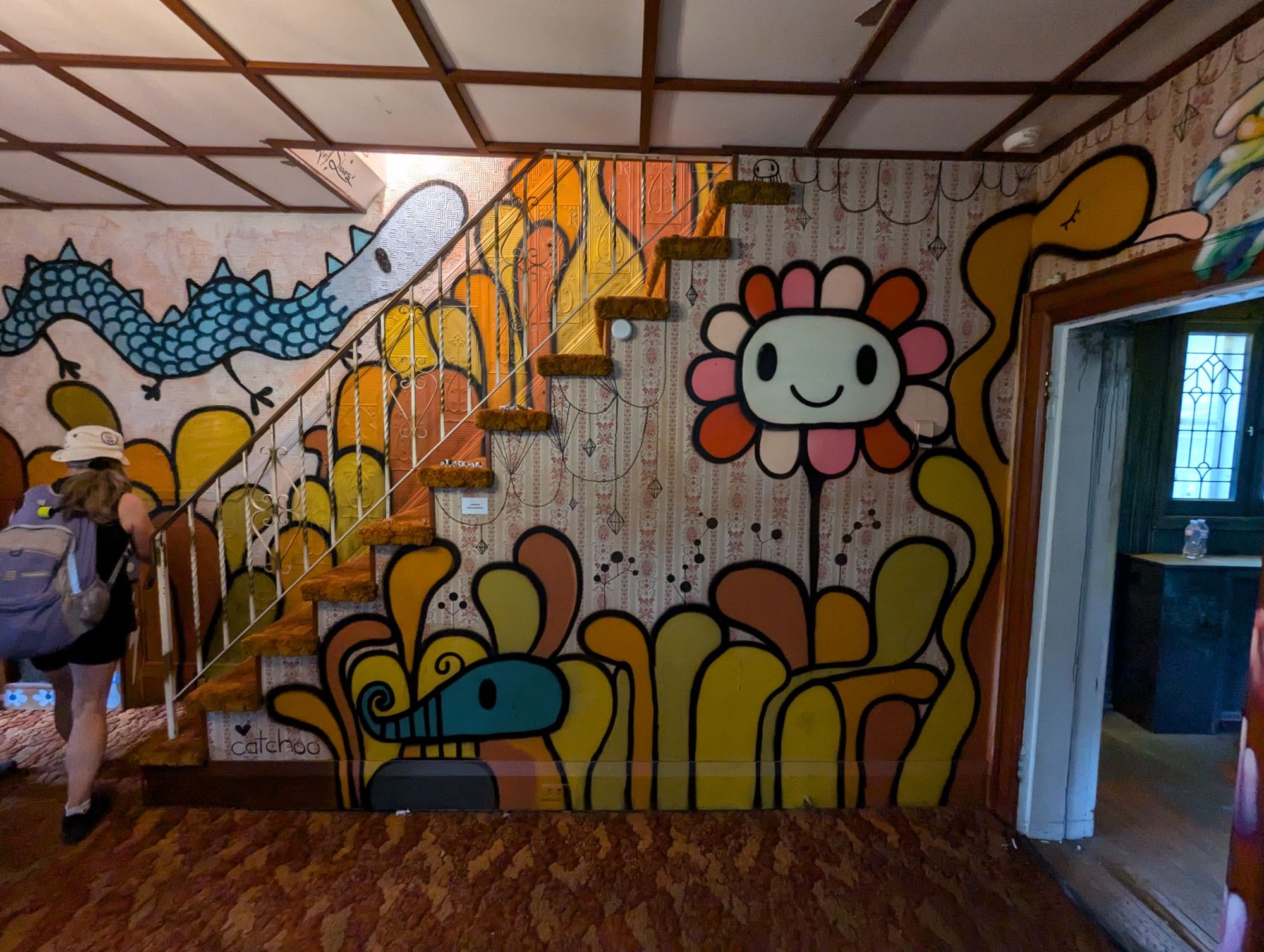 On a wall with a staircase in it, a bunch of cartoon wiggly yellow and orange plants, a teal but, a pink petalled flower, and a long snub nosed blue scaled dragon. The room has brown 70s carpeting