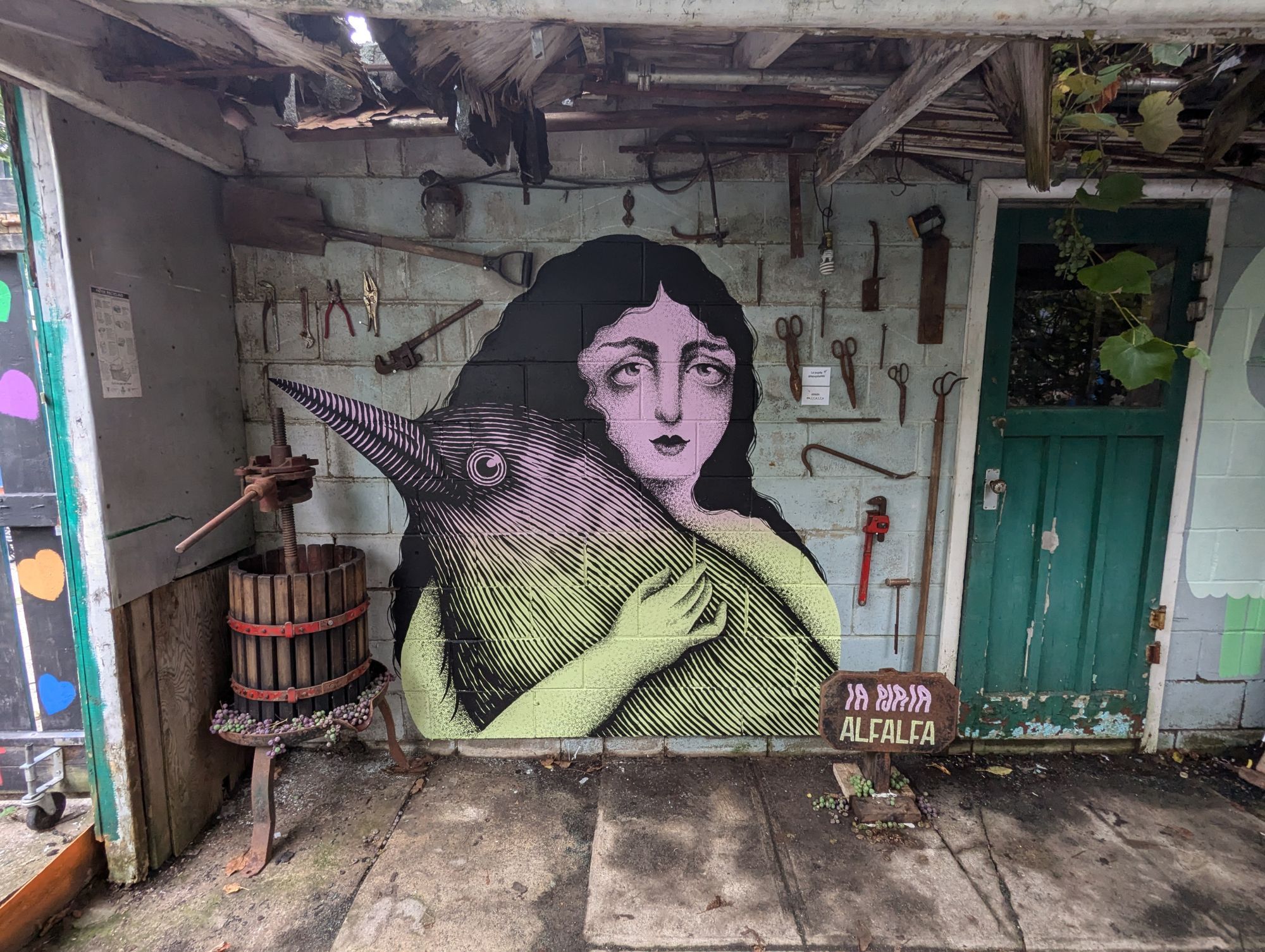 A back shed with the bust of a woman holding an enormous bird, painted with large black areas and thing lines, almost like scratchboard, with pale yellow green fading to pink as a base colour. The pale green wall has tools hanging on it, such as a shovel, crowbar, monkey wrench, etc.