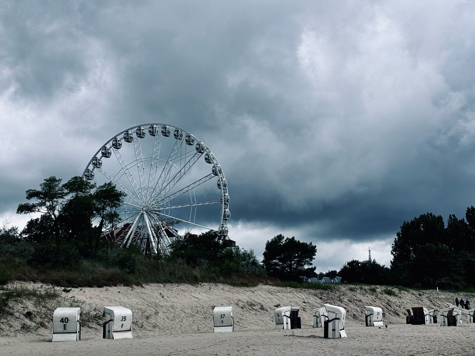 Das Bild zeigt einen Strand mit mehreren weißen Strandkörben, die in einer Reihe auf dem Sand stehen. Im Hintergrund erhebt sich ein großes Riesenrad, das von Bäumen umgeben ist. Der Himmel ist mit dichten, sehr dunklen Wolken bedeckt, was auf bevorstehendes oder aktuelles schlechtes Wetter hindeutet.