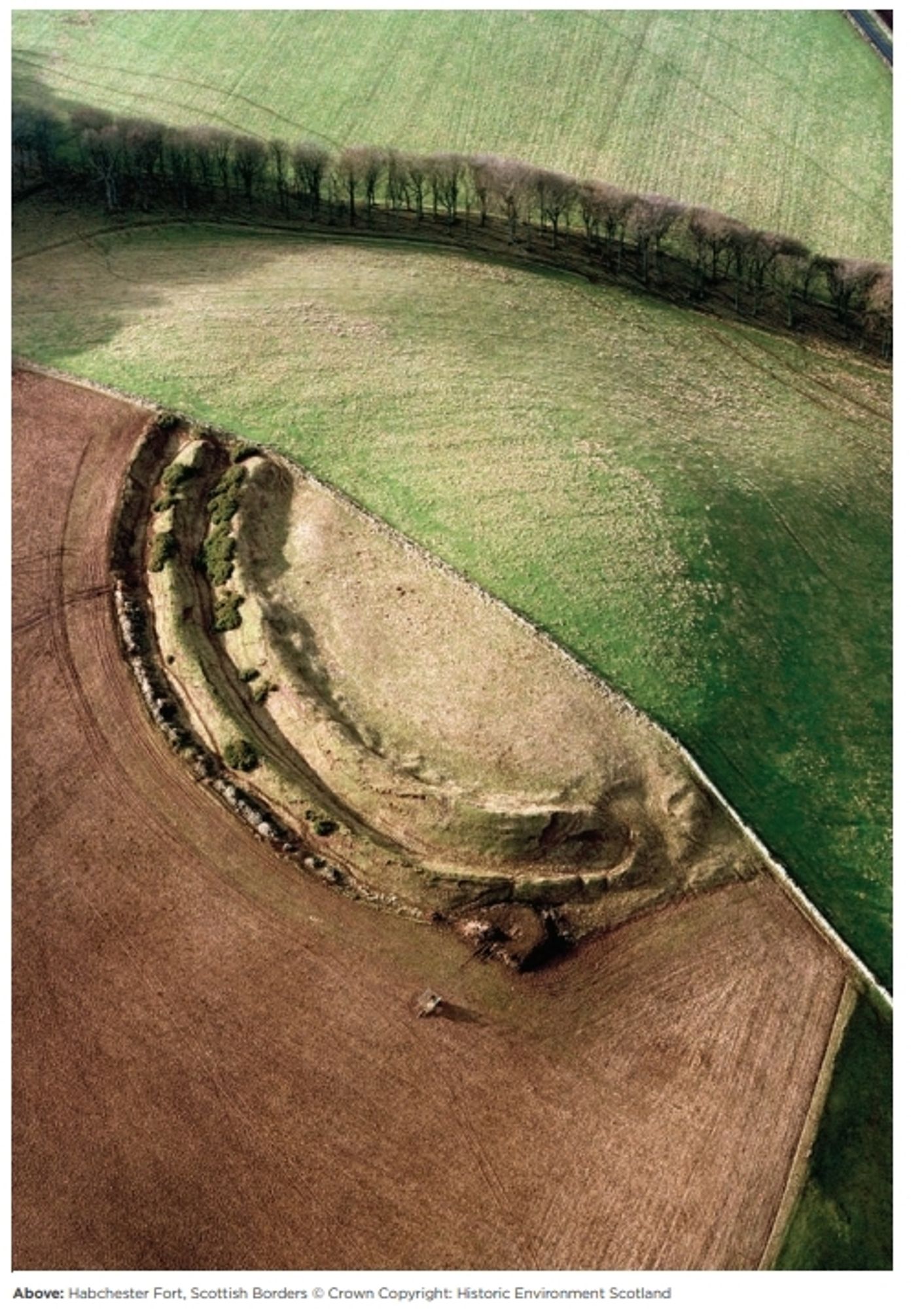 aerial view of an earthwork considting of two banks ans ditches. the monument is divided in half by a field boundary. on one side it is well preserved, on the other ploughing has destroyed the structure which is visible only as a faint trace