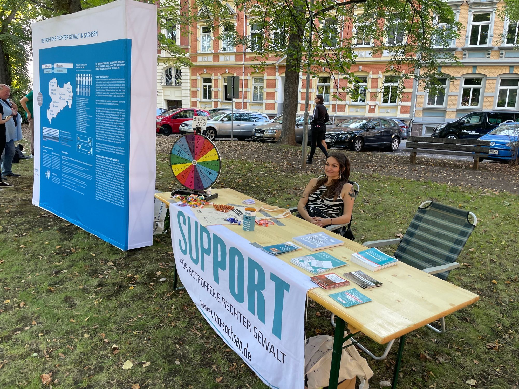 Bild vom Infostand mit Flyern unserer Projekte sowie einem Glücksrad und einem Banner mit dem Logo von Support. Links im Bild sieht man eine Infowand mit Zahlen zu Betroffenen rechter Gewalt