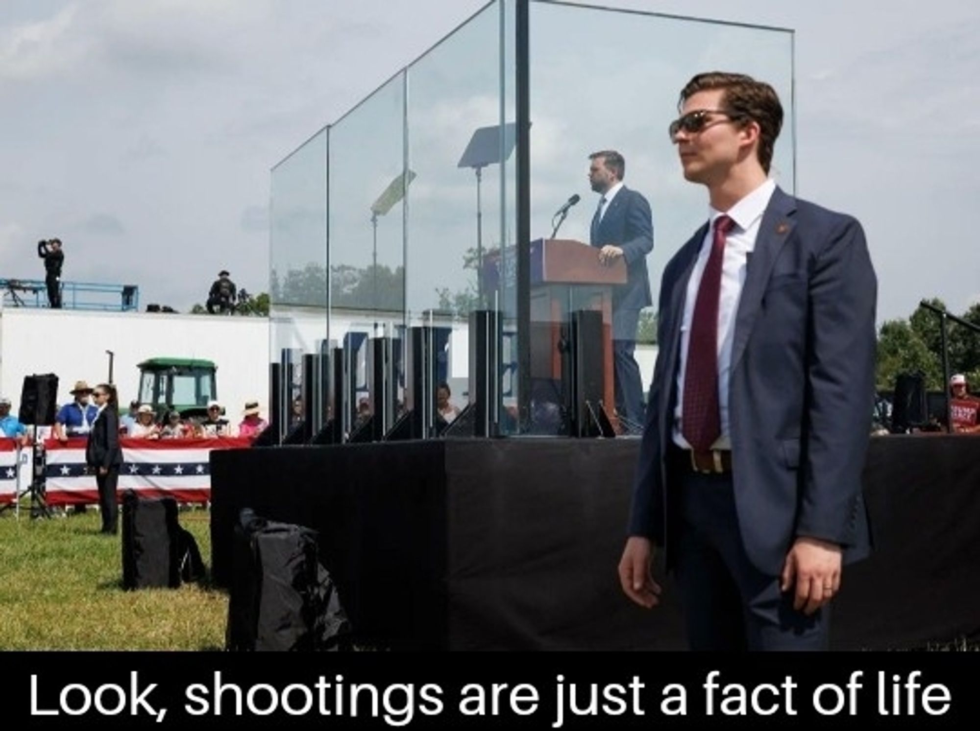 JD Vance on stage behind bulletproof glass with guards posted at either side 
Caption: Look, shootings are just a fact of life