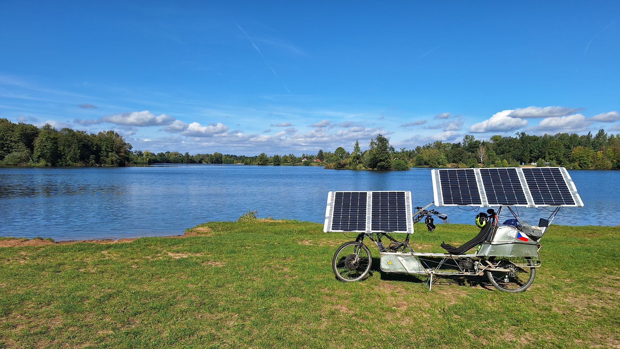 Solar bicycle parked near the lake. Blue sky with distant little clouds on the horizon, green grass, blue walter, green trees.