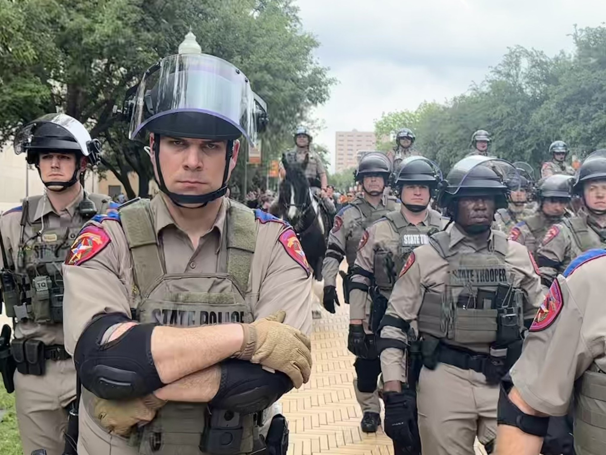 A bunch of Texas DPS State Troopers prepare to invade The University of Texas. There's a cop in the front with his arms crossed. He's trying very hard to look mean, but he really just looks like a dick.