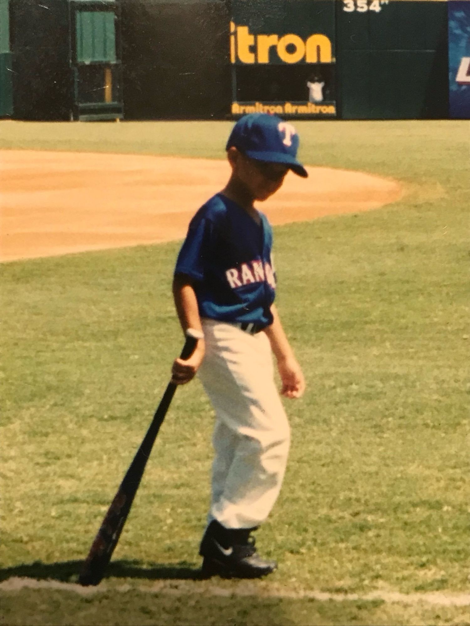 Patrick Mahomes as a child, dressed in his full bat boy gear at the Ballpark in Arlington. He’s walking along the baseline, wearing a blue Texas Rangers jersey, oversized white baseball pants and black cleats. He’s dragging a large wooden baseball bat behind him and looking slyly to his right, towards the camera