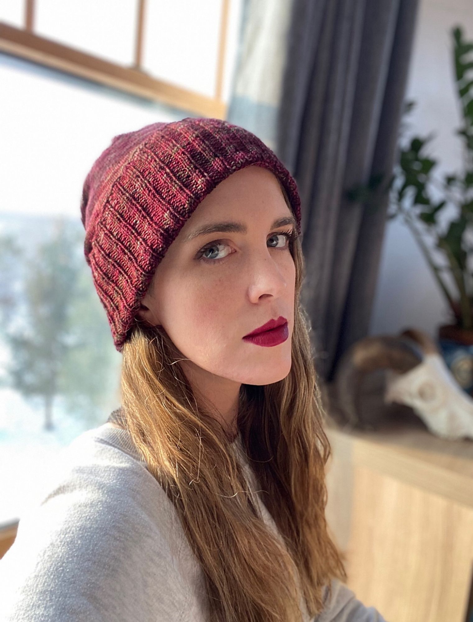 The knitter modeling a hand-knit hat in a dark red variegated yarn. The hat perfectly matches her lipstick. There is a ram skull in the background that briefly achieved minor fame on #uglydogs Twitter.