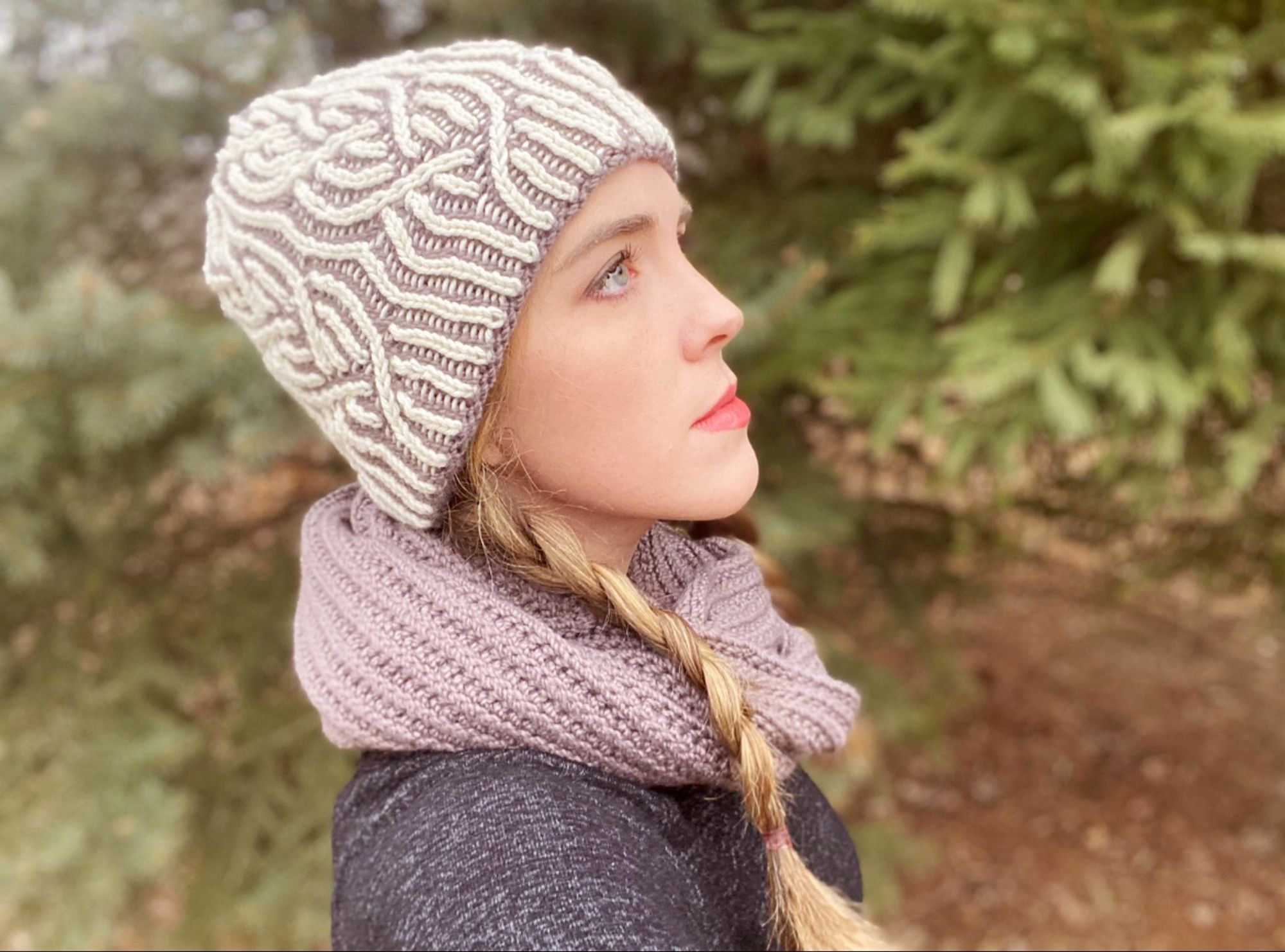 The knitter modeling a hand-knit taupe and white hat, featuring a pretty twisted brioche design. She is also wearing a hand-knit infinity scarf in the same taupe yarn has the hat.