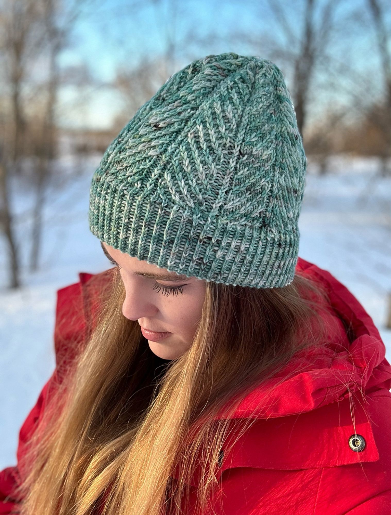 The knitter modeling a hand-knit hat in a custom dyed variegated sea green yarn, featuring an unusual diagonal rib construction. She is wearing a bright red parka in front of a snowy background.