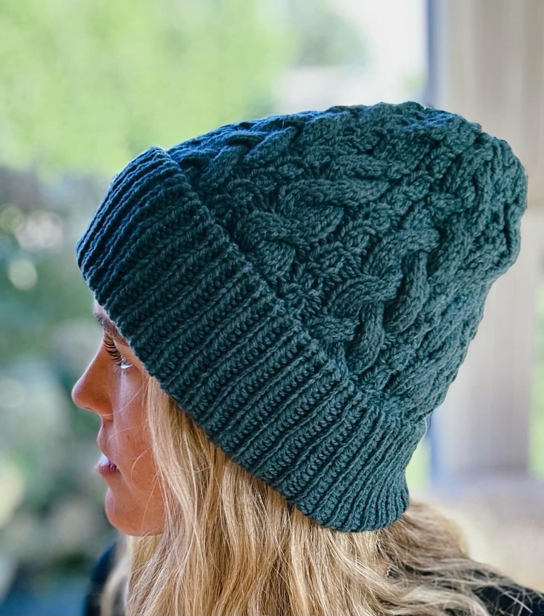 The knitter modeling a heavily cabled hand-knit hat in a moody forest green yarn.