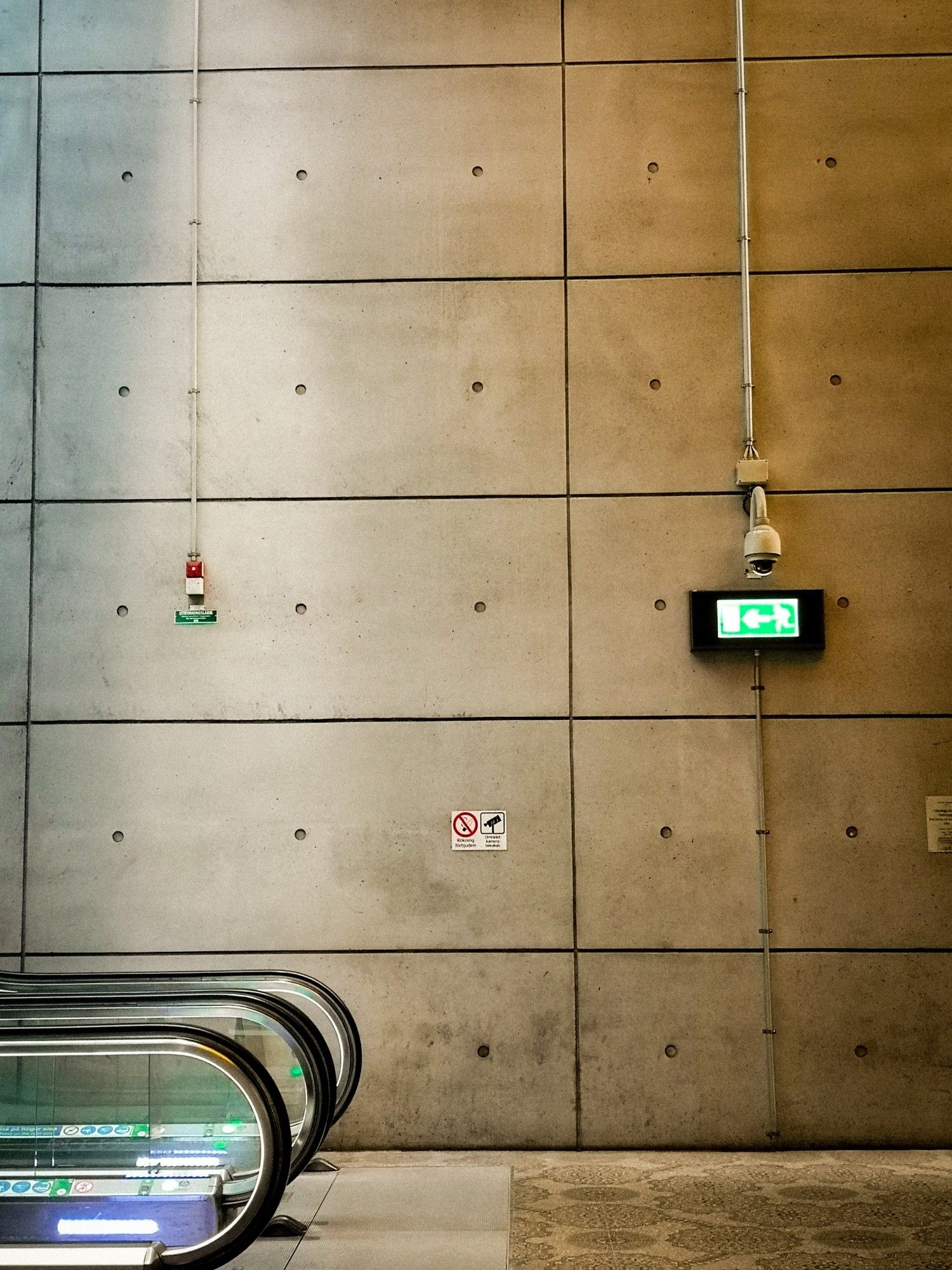 Links unten Rolltreppen zu sehen.
Im Hintergrund Wand aus Beton. Rechts mittlere Linie ein "Notausgang" Schild.