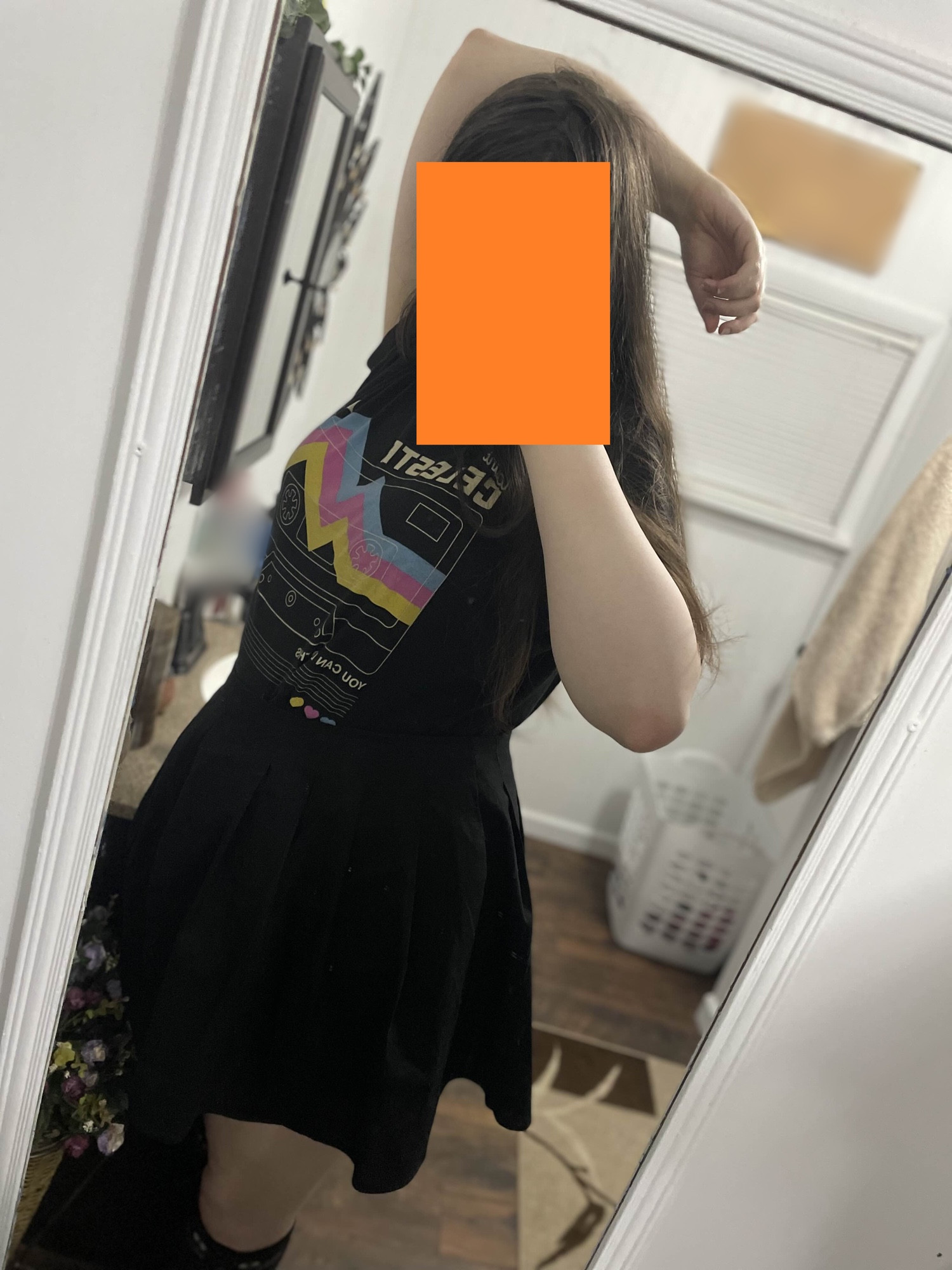 A picture of Wonder IRL, she has pale white skin with long dark brown hair, her right arm is resting on top of her head, while she takes the picture in a mirror with her left arm, she is slightly turned to her right. She is wearing a black shirt based off of a Celeste Cassette that is black, with blue, pink, and yellow streaks indicating a mountain, with the text Celeste and You Can Do It on it. She is also wearing a black skirt with black cat knee socks.