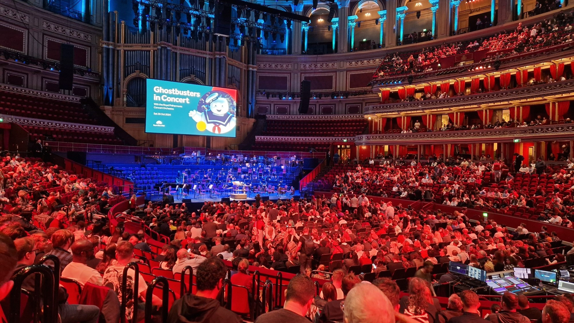 A picture of the Royal Albert Hall before a concert. It is lit in reddish tones for the audience, and blue tones for the stage.