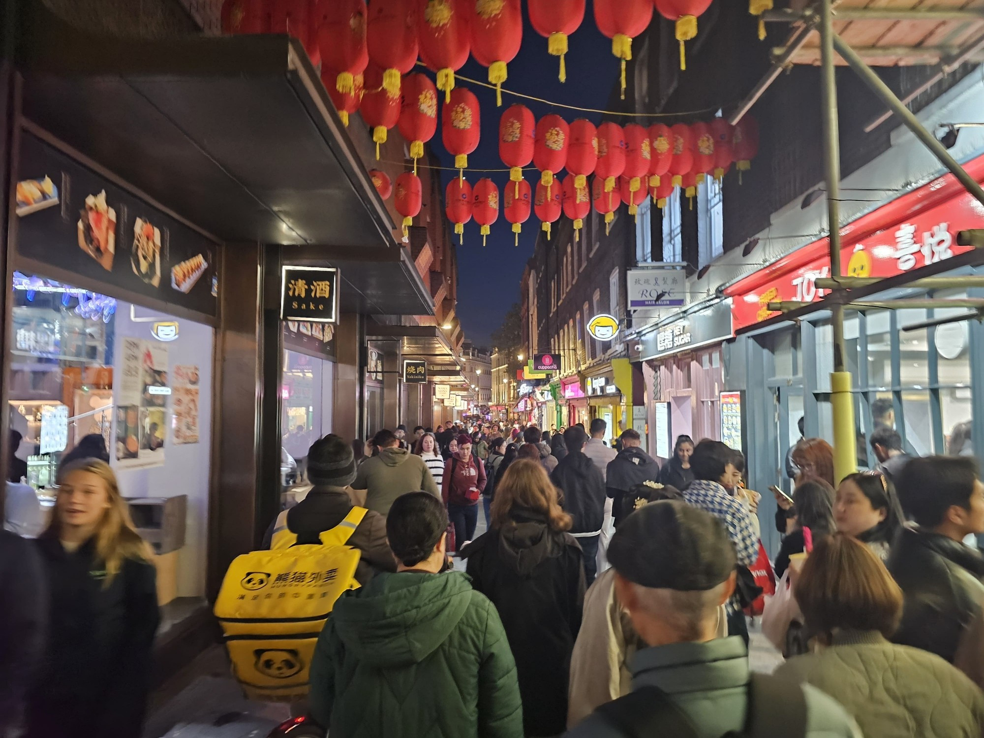 A picture of a street in Londons China town in the evening. Red lanterns hand over head. The street is full of people