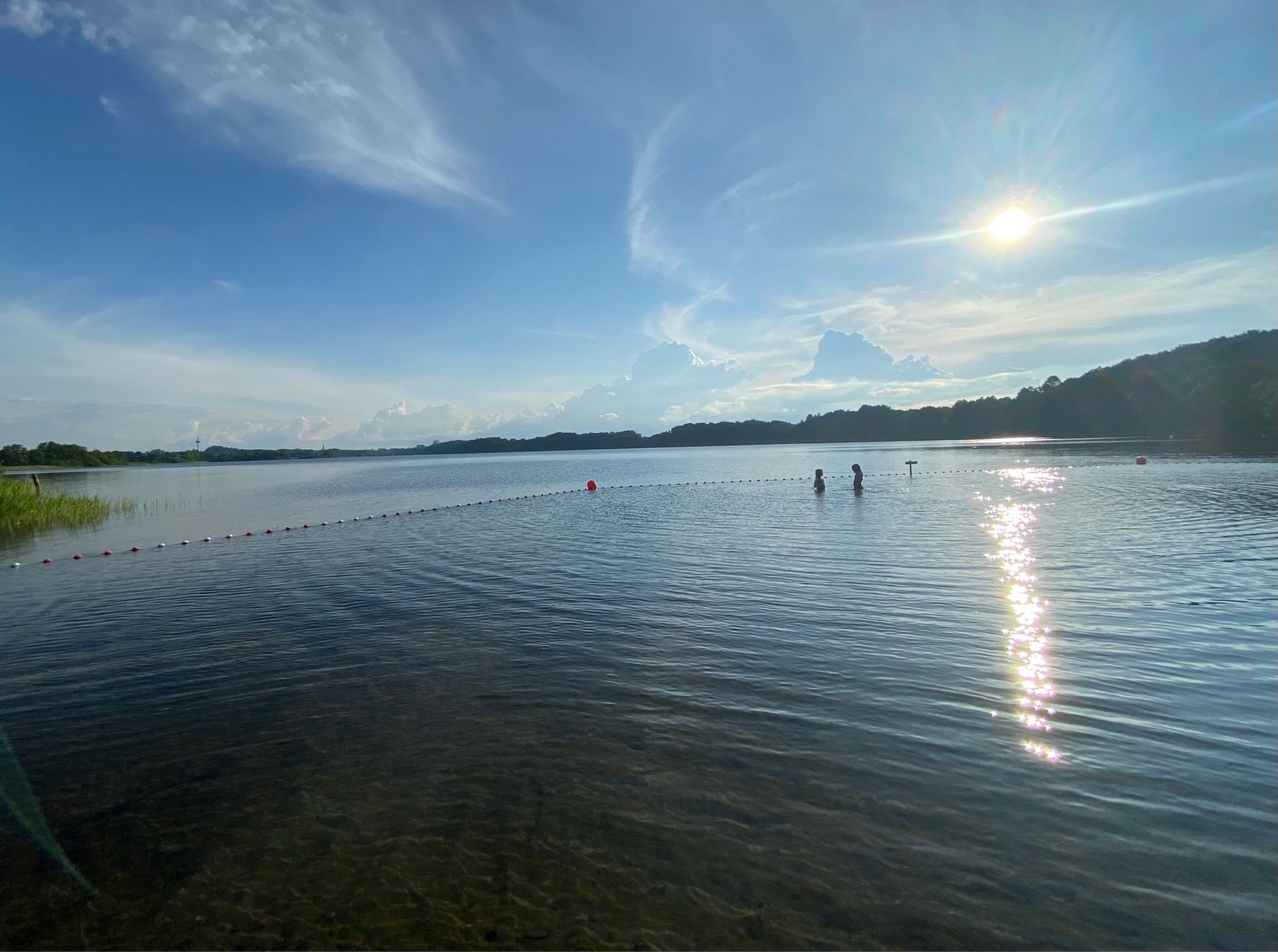 Weiter Blick über den See, Gegenlicht, weit hinten ganz klein Baumreihen, darüber blauer Himmel mit einigen Wolken