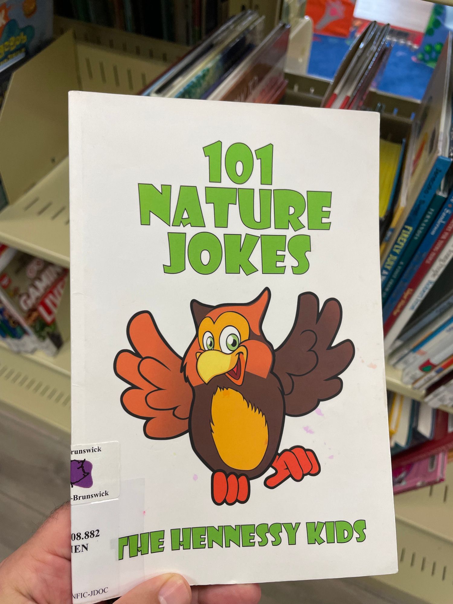 A copy of 101 Nature Jokes by The Hennessy Kids held in hand, a smiling cartoon owl on the cover. In the background are the library stacks in the children's section of a public library.