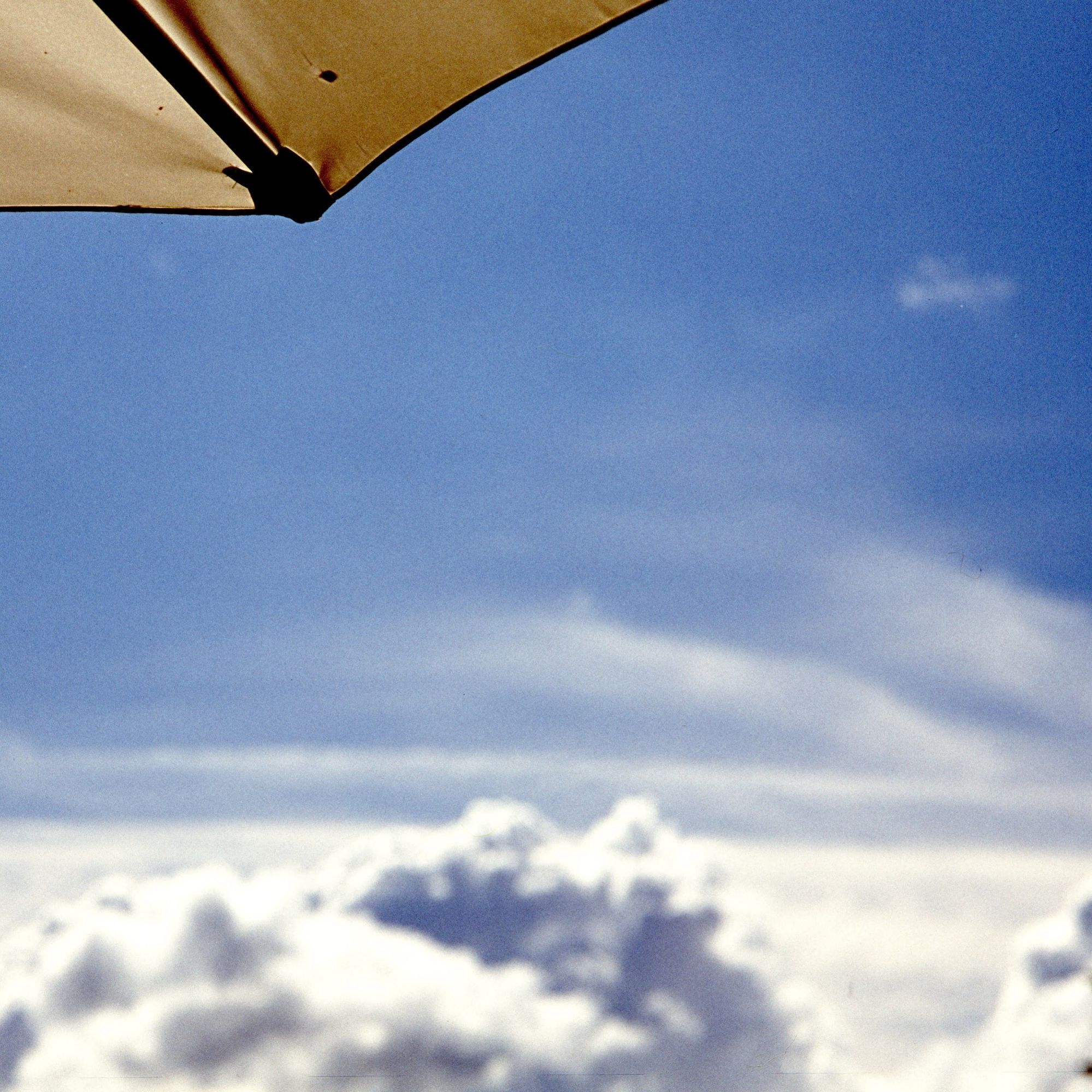 Drei Viertel blauer Himmel, am unteren Rand ein paar weiße Wolken in Unschärfe, oben links ragt ein Stück gelber Sonnenschirm mit einer schwarzen Strebe ins Bild.