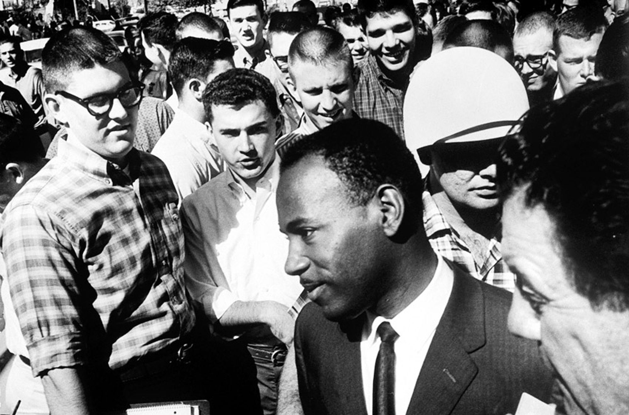 A picture from the Ole Miss Riot from 1962 where a white crowd protested violently against desegregation of the University of Mississippi. Black student and veteran James Meredith in a well fitting suit and tie is walking by a crowd of white protesters in casual clothing. James Meredith has a calm expression, while the white men in the crowd look at him with disgust and mockery.