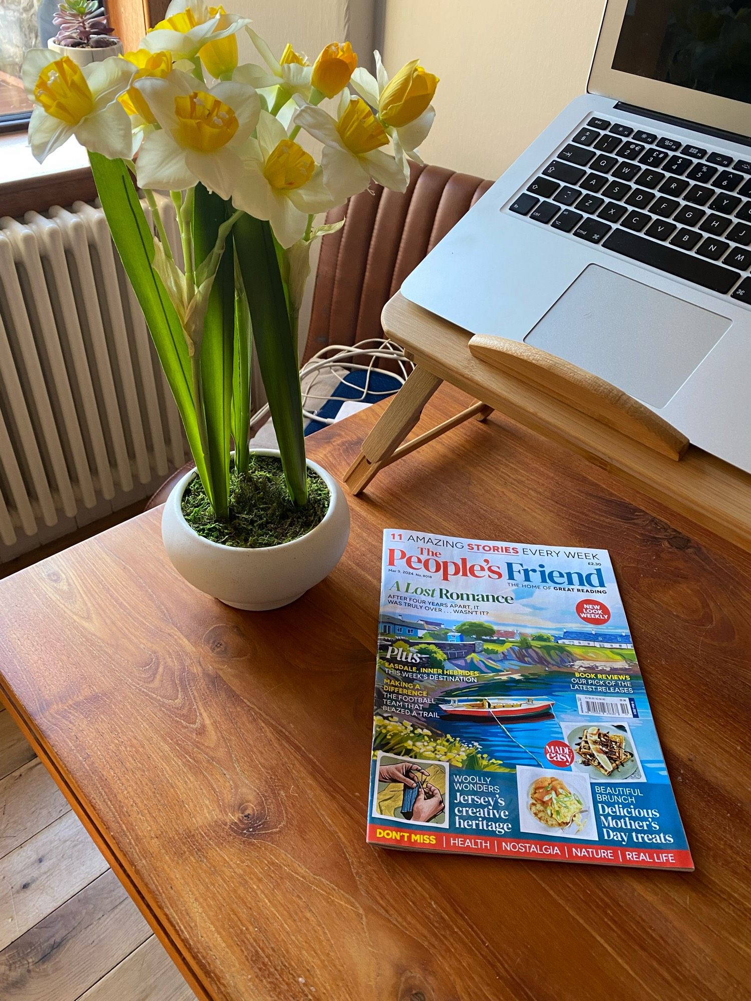 Wooden table holds a pot of tete e tete daffodils, a People’s Friend magazine and a laptop.