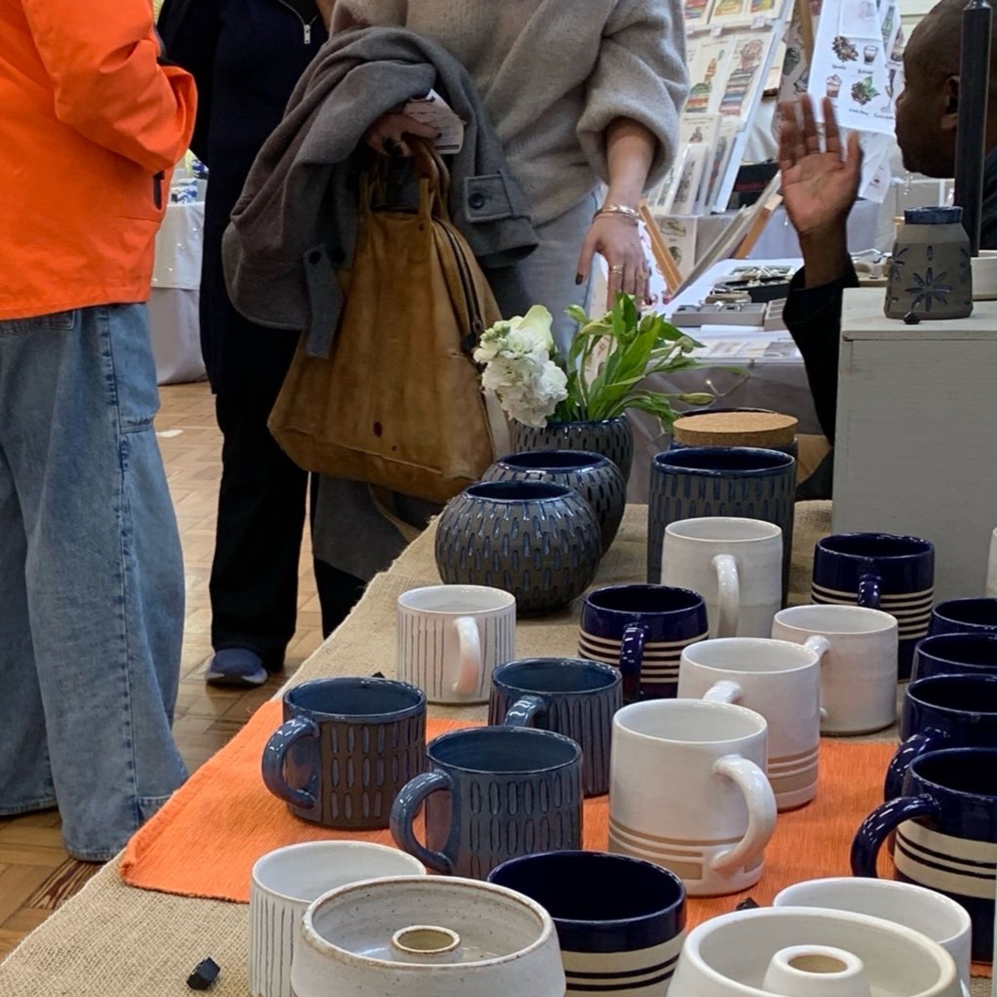 A table full of pots at a market. Many are striped, all are neutral colours or blue. There are people in the background chatting.