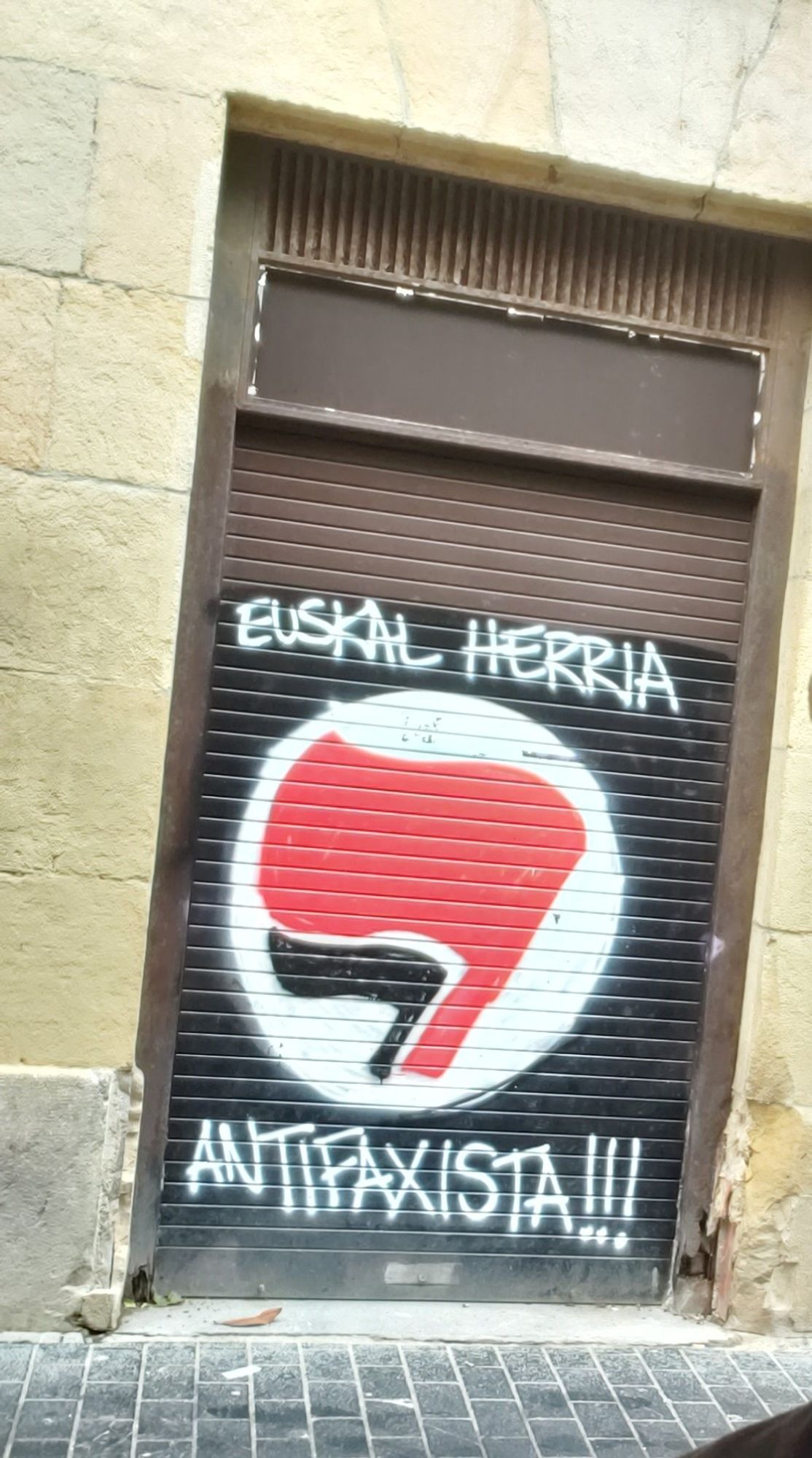 A closed metal shop gate spray painted with red and black flags against a white background and text reading "Euskal Herria Antifaxista!!!" 