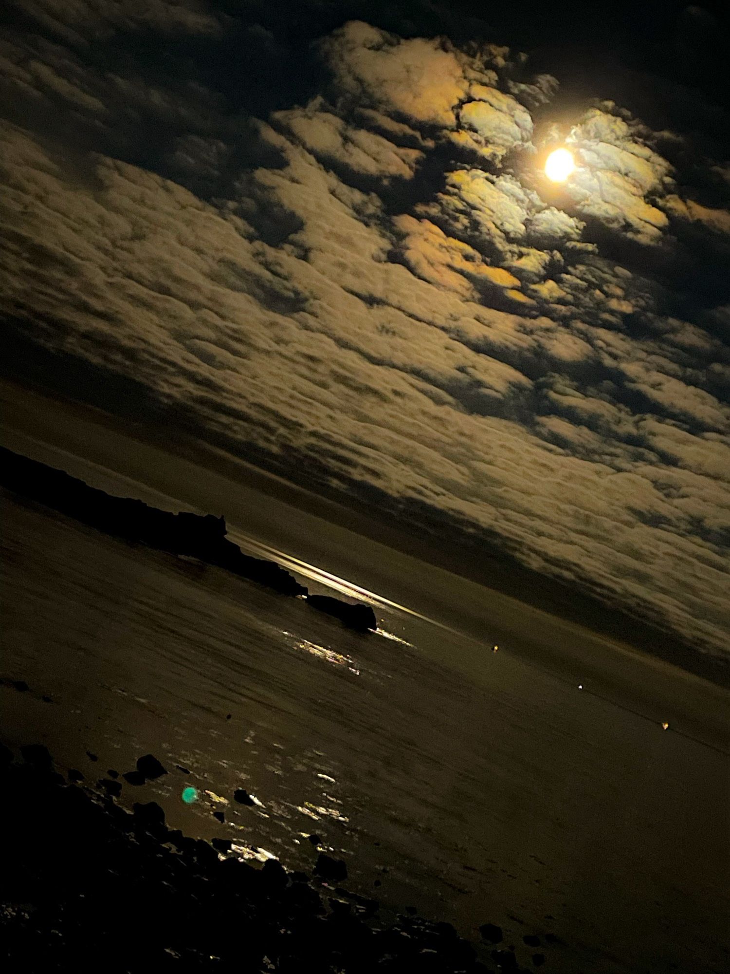 Golden yellow full moon shining through dappled clouds and reflecting on the flat surface of the sea around a small rocky islet