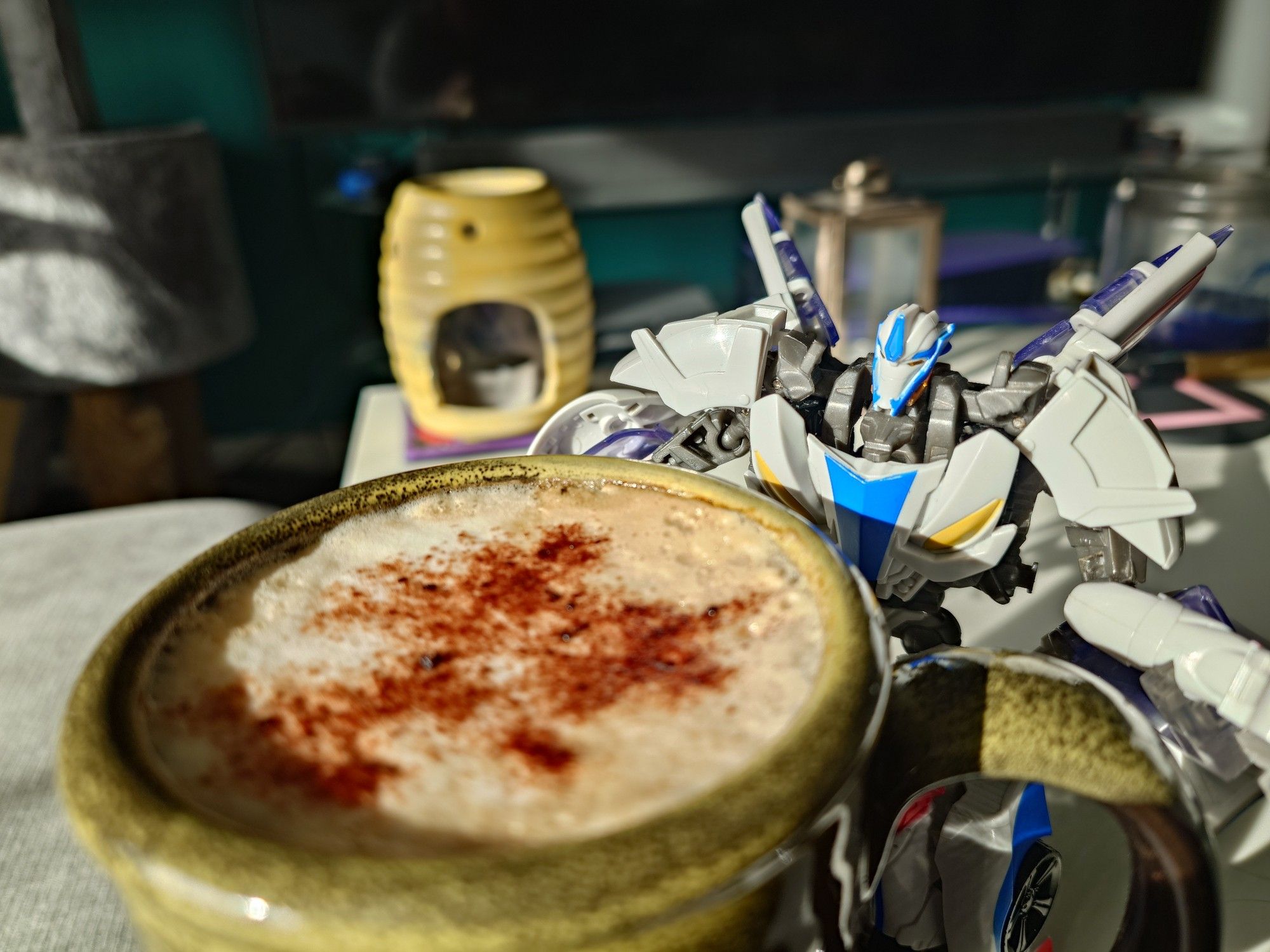 A Transformers Prime Smokescreen figure stood behind a large cup of foamy coffee with chocolate dusted on top. The morning sun shines on the image.