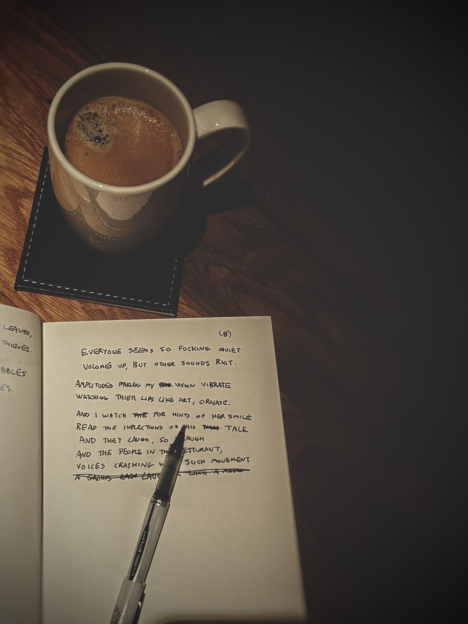 A top down view of coffee on a coaster and an open notebook with an unfinished poem that says

EVERYONE SEEMS SO FUCKING QUIET.
VOLUMES UP, BUT OTHER SOUNDS RIOT.
AMPLITUDES MAKING MY VISION VIBRATE.
WATCHING THIER LIPS LIKE ART, ORNATE.
AND I WATCH FOR HINTS OF HER SMILE
READ THE INFLECTIONS OF HIS TALE.
AND THEY LAUGH, SO I LAUGH.
AND THE PEOPLE IN THE RESTAURANT
VOICES CRASHING WITH SUCH MOVEMENT