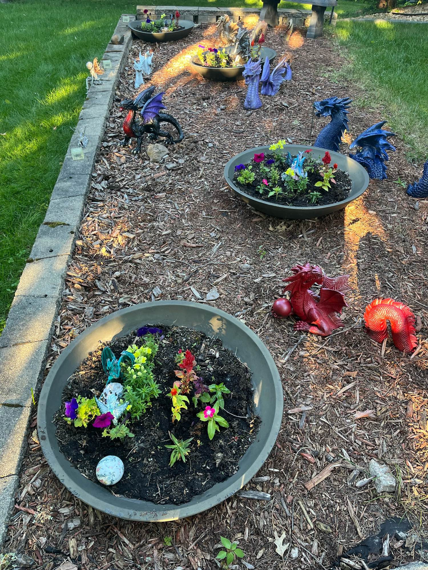 Four large, dark grey flower bowls each with a fairie and small dragon. Two fairies sitting on the left side of the flowerbed wall. A red, blue, purple, gold, white, and multi colored dragon on mulch in the bed. The multi colored dragon is black with purple wings and red on its neck from its chest to its head.