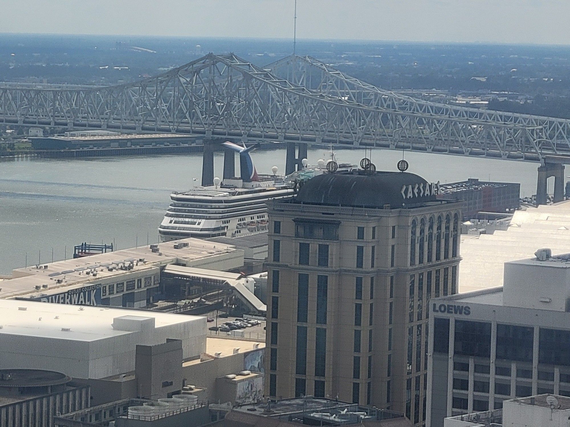 A huge cruise ship in the Mississippi River in New Orleans, with city buildings in the front and a large bridge behind.