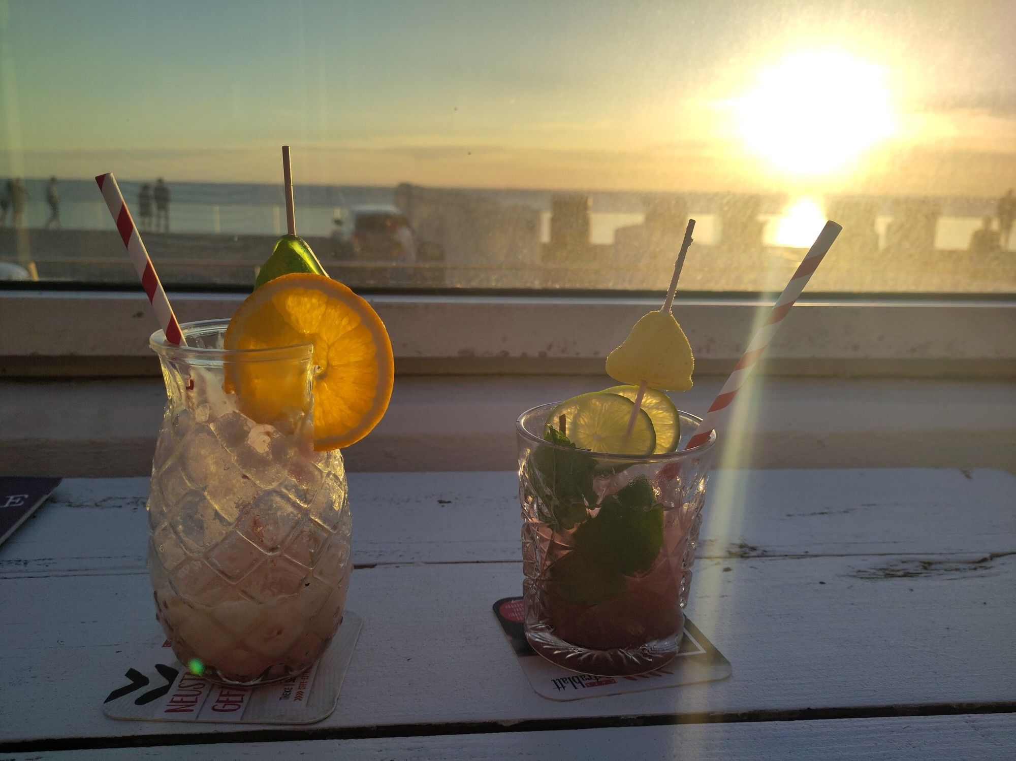 Auf einem Tisch im Gegenlicht bei untergehender Sonne mit Strandkörben und Meer im Hintergrund stehen zwei Cocktail Gläser. Eis ist fast leer. Das andere fast voll.