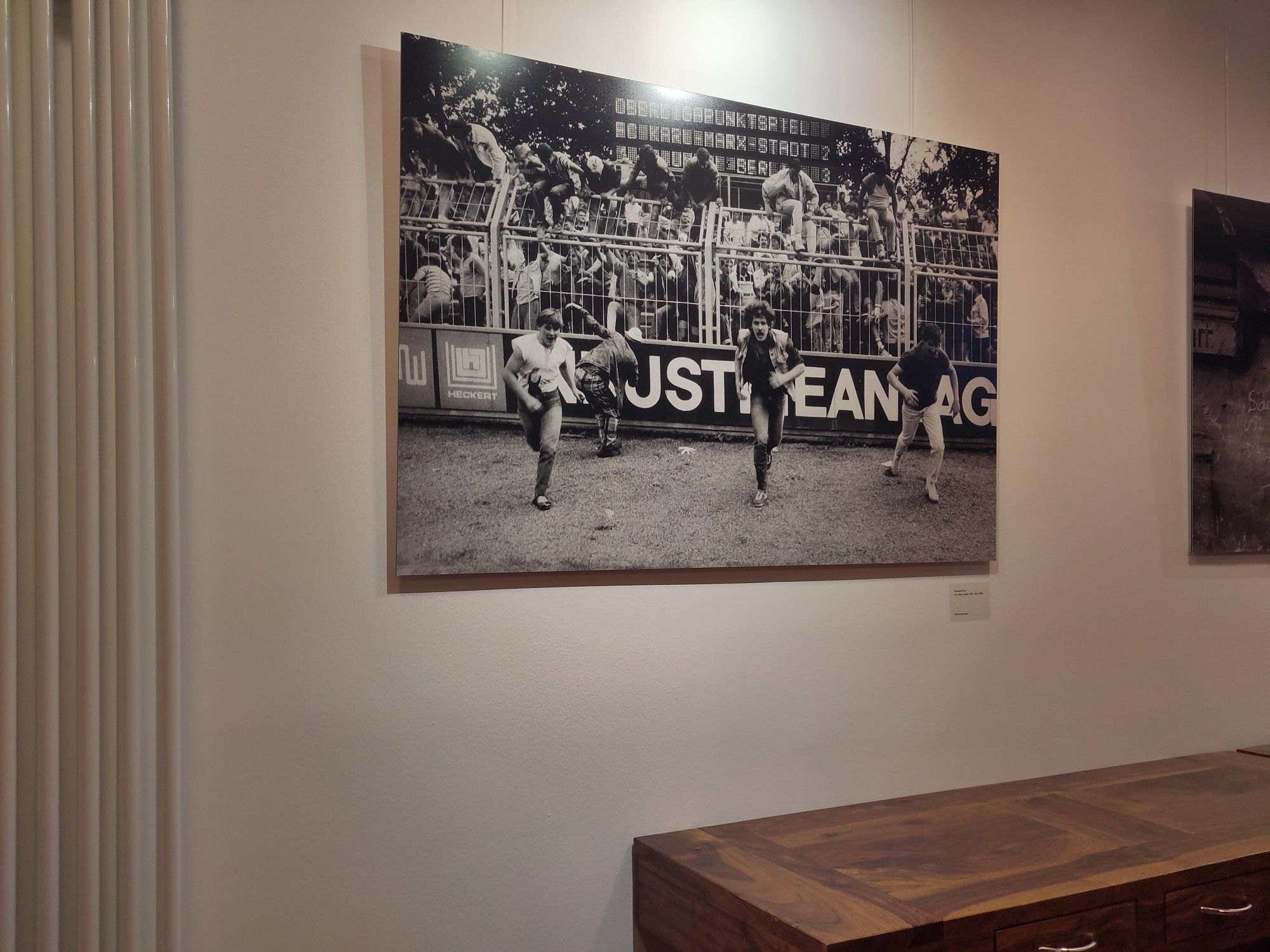 Ein Bild in einer Ausstellung. Schwarz Weiß. Fußballfans klettern über den Zaun auf den Rasen.