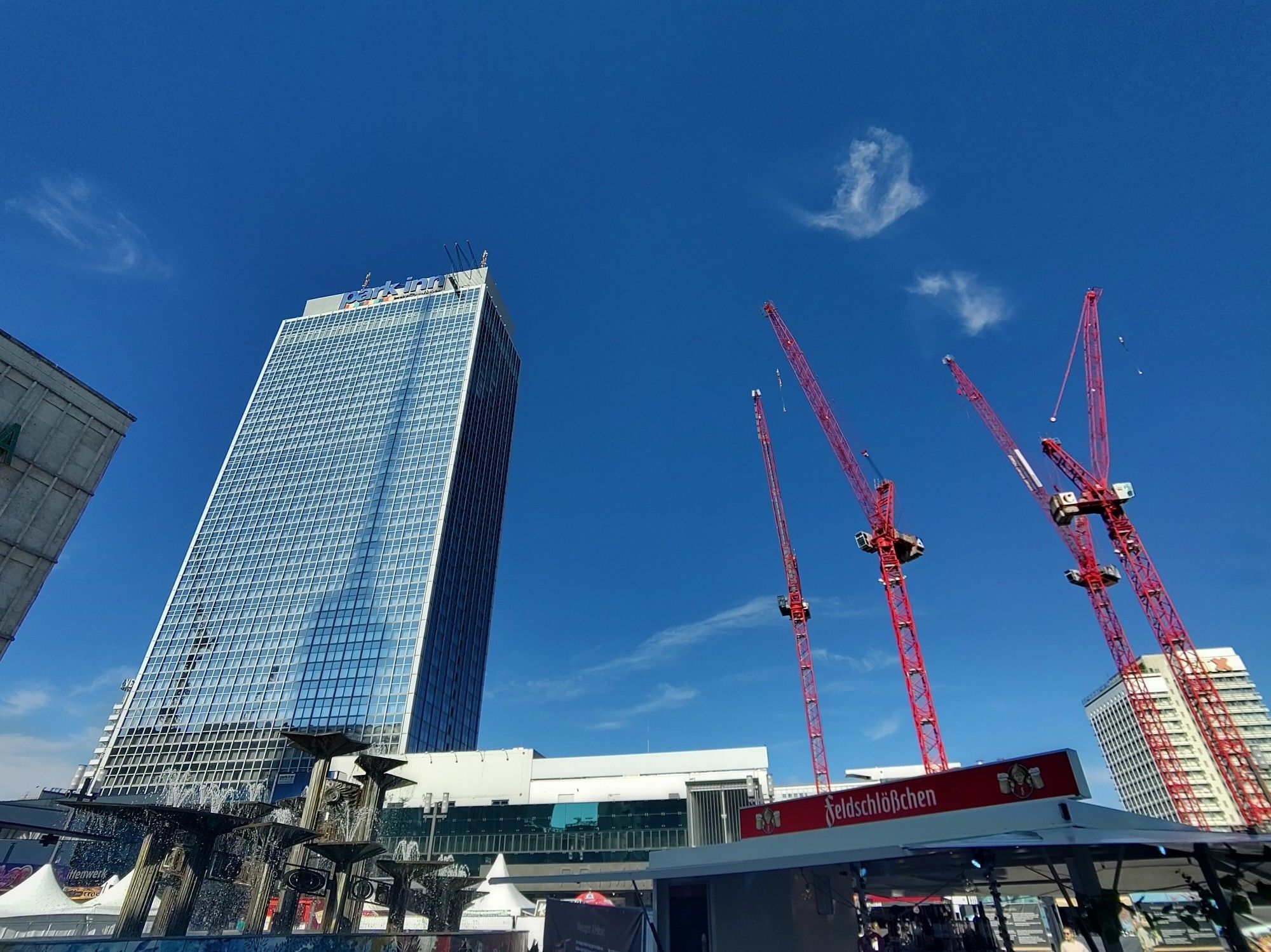 ParkInn am Alexanderplatz. Kräne, Brunnen, Blauer Himmel, Menschen, Buden. Im ParkInn ist der Schatten des Fernsehturm zu sehen.