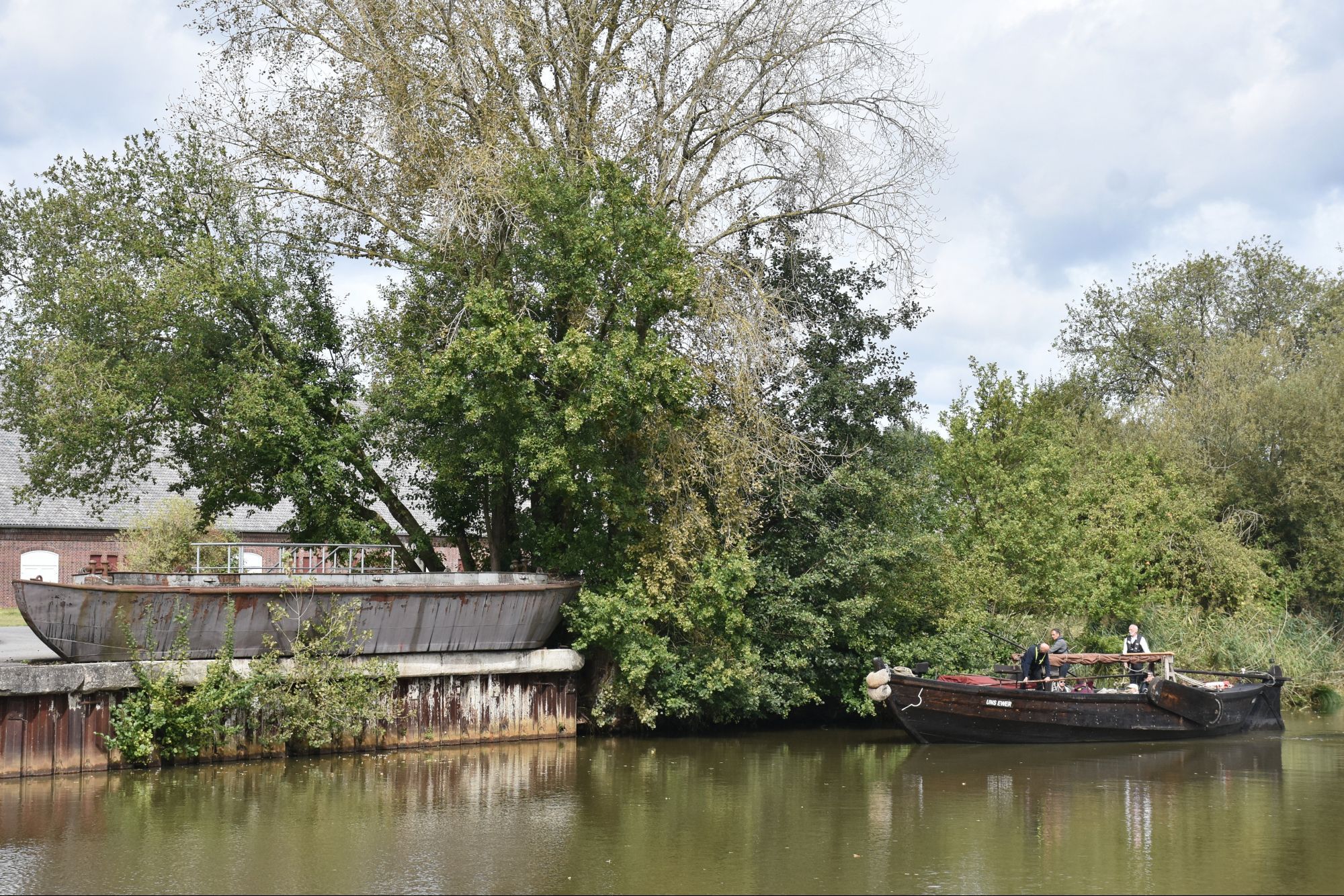 Das historische Frachtschiff, ein Ewer, fährt auf der grünfarbigen Dove-Elbe. Im Hintergrund viele grüne Bäume. Der Ewer ist kurz vor einer Spundwand. An Land steht ein alter Lastkahn. Foto: SHGL