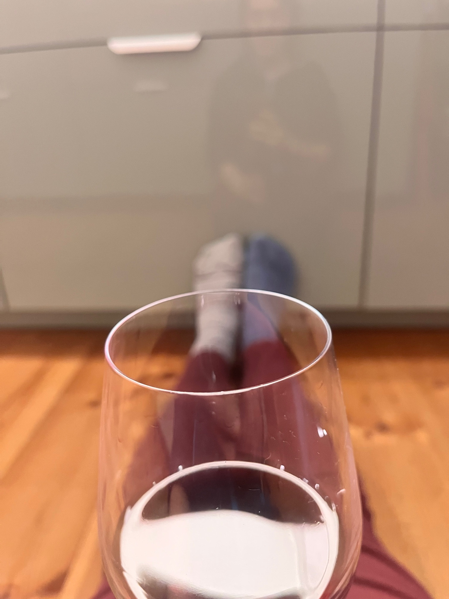 Foto aus meiner Perspektive. Ich sitze auf dem Fußboden. Im unteren Bereich des Fotos ist ein Glas Wein. Im Hintergrund sind unscharf meine Beine und Füße, die an einer Küchenfront lehnen.