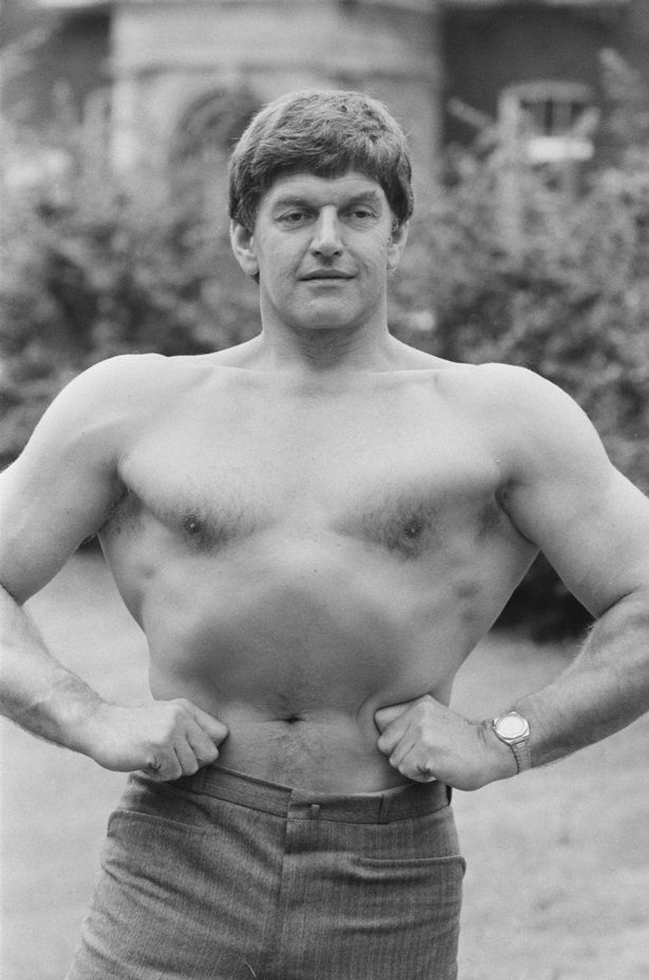 Bw image of a shirtless, somehow extremely obviously British man, flexing his lats. His body is hairless, his pec and upper trap development is lacking compared to modern aesthettics, he is notably muscular but not dehydrated and shredded.