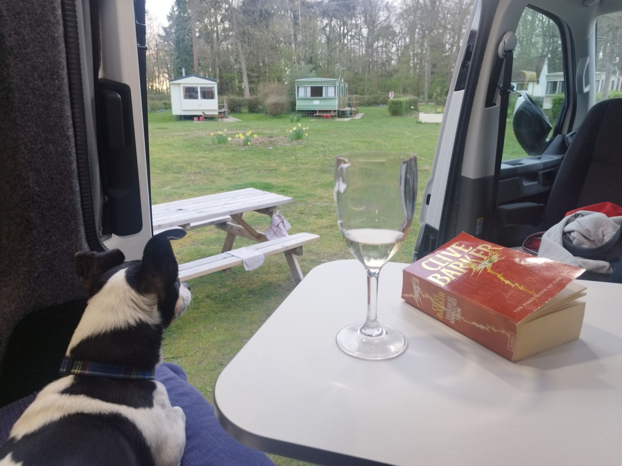 The view from inside a campervan. A sunny campsite. Inside is a Jack Russell dog, a glass of wine and a paperback copy of The Great and Secret Show by Clive Barker.