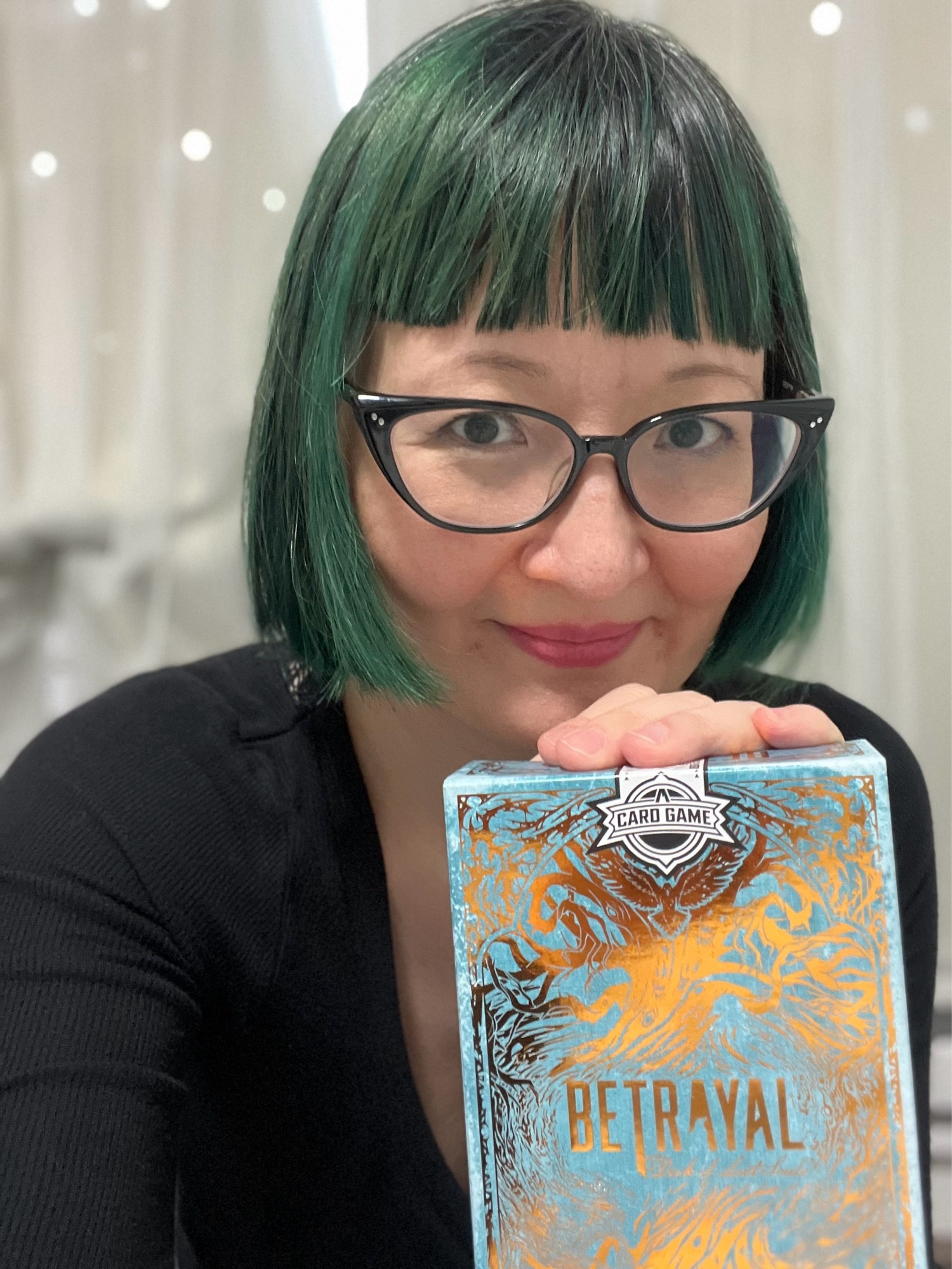 It’s me, Kat Kruger, with straight bangs and a short green bob haircut. I’m wearing black cats eye classes and have a hand on top of a gold foil box of Betrayal Deck of Lost Souls.