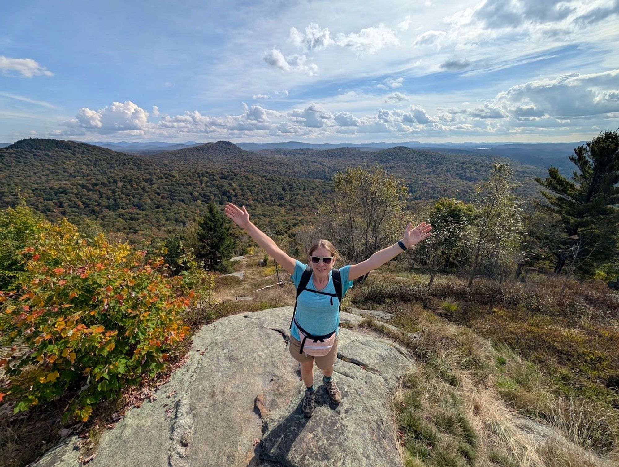 A photo of me standing on the summit of Goodman Mountain. I'm wearing a green hiking shirt, tan shorts, a pink fanny pack, a black pack, and brown hiking boots. My light brown hair is tied back, and I wear green sunglasses. I raise my arms in joy and triumph while green mountains spread out in the distance. Autumnal colors pop here and there under a blue sky dotted with white clouds.