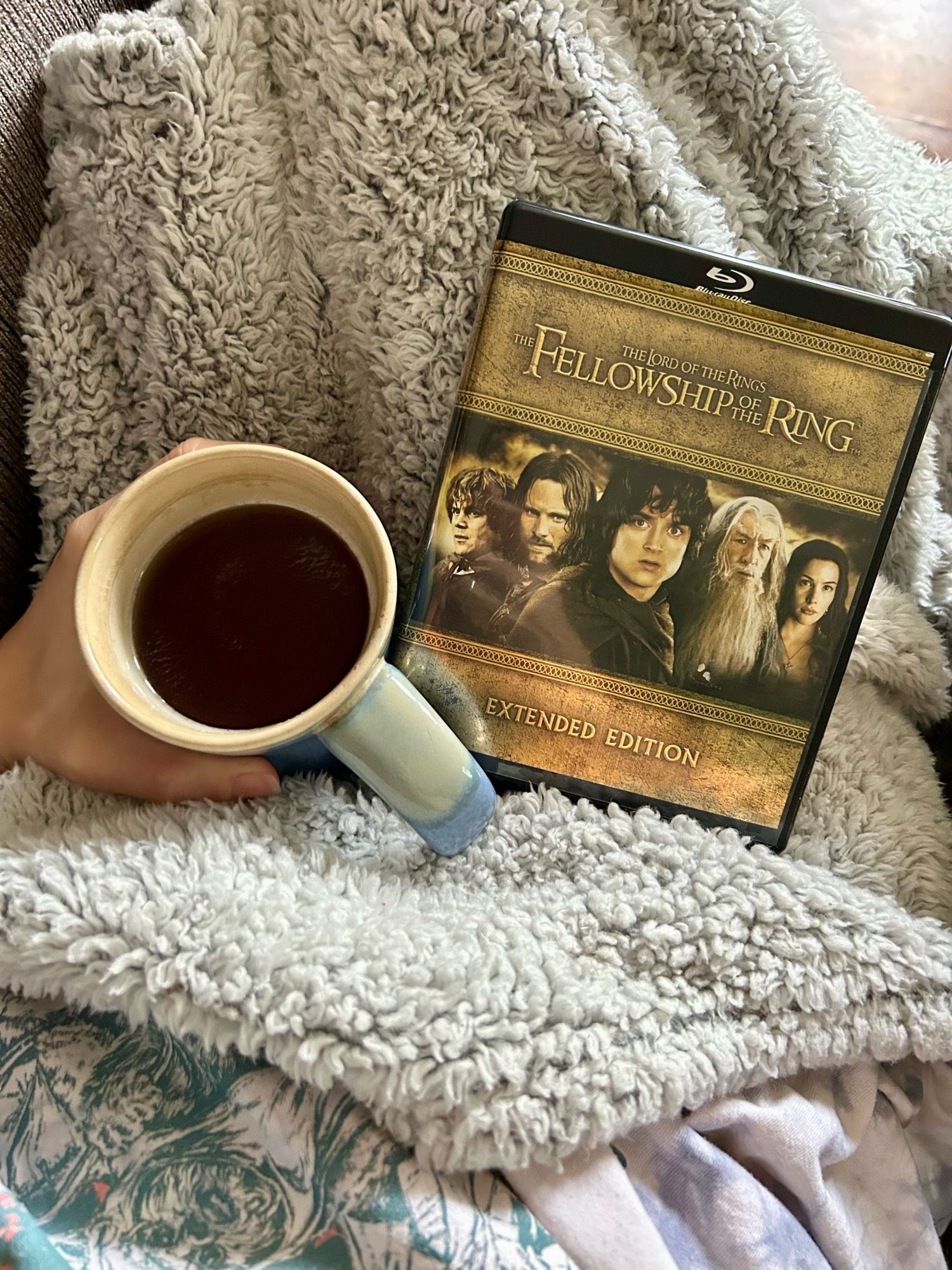 A hand holding a full mug of tea beside the case of the extended edition of Fellowship of the Ring. Everything rests on a fluffy gray blanket.