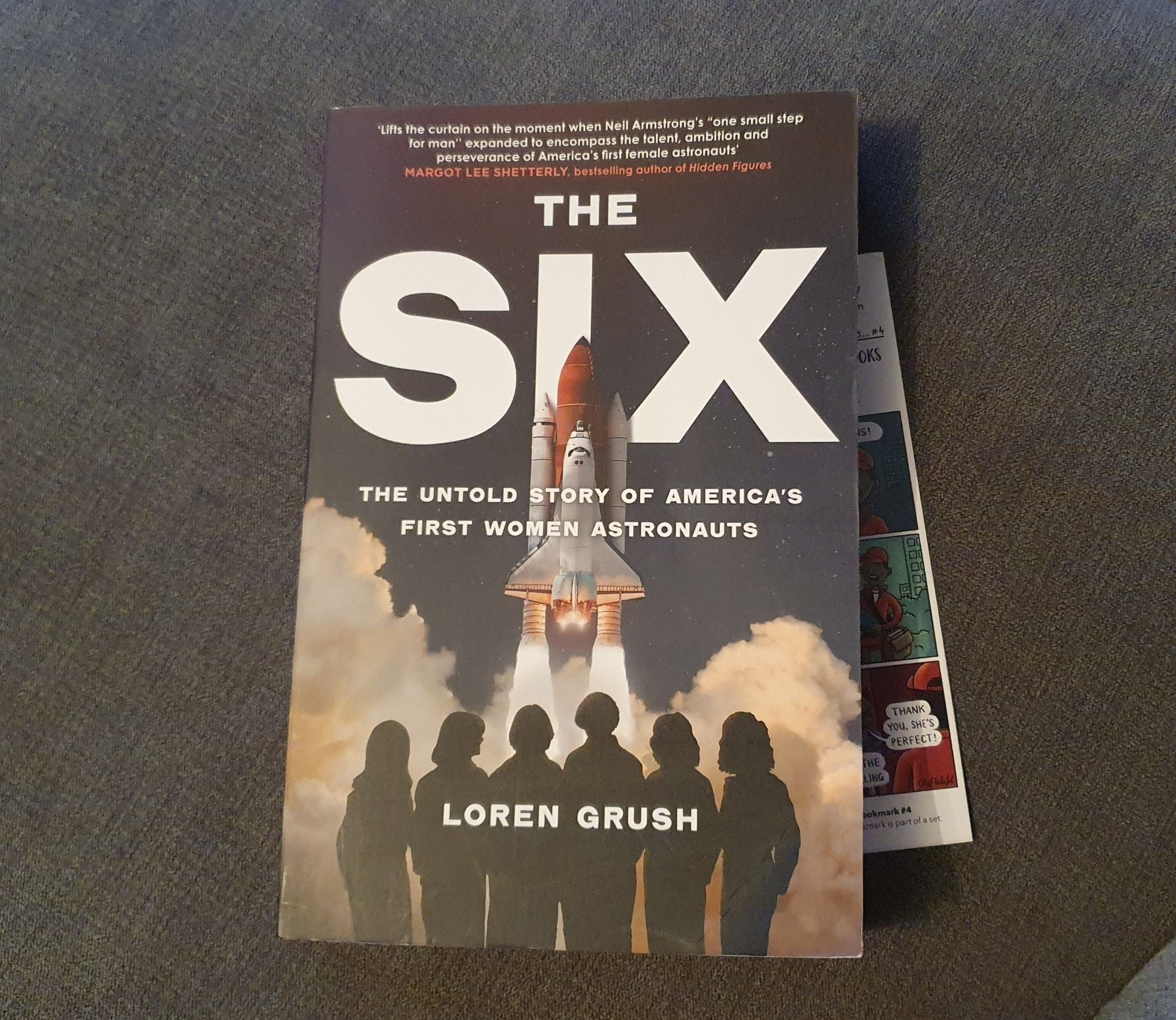 Paperback copy of The Six by Loren Grush. Bookmark visible to the right