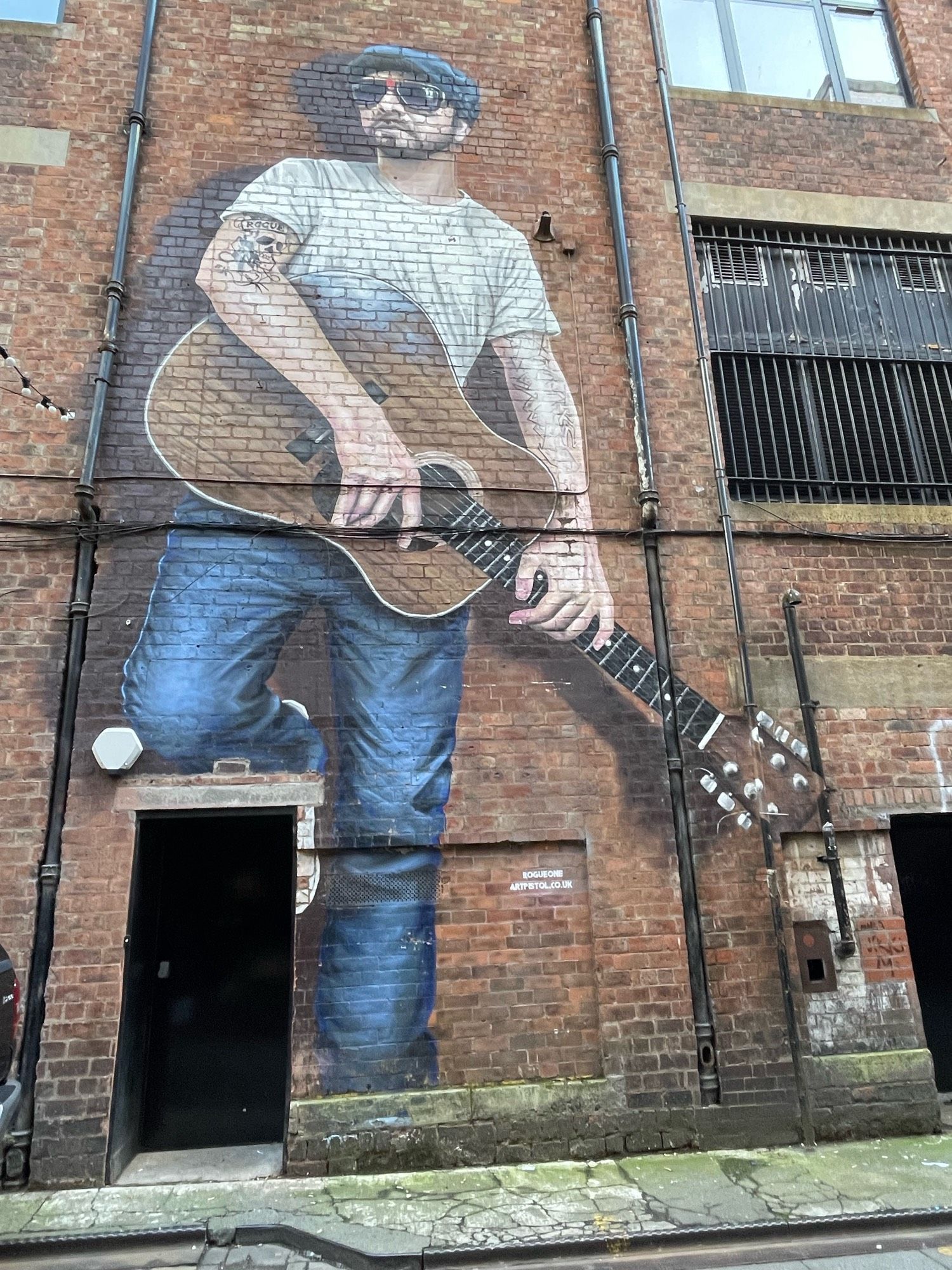 A mural on a brick wall on Sauchiehall Lane in Glasgow. It is mainly between two downpipes and is of a man with an acoustic guitar, in a white t shirt and blue jeans - his right knee is resting on the top of a doorframe. He has a small trimmed beard and is wearing a cloth cap and aviator sunglasses. He has a tattoo with a skull on his right bicep with the wording ROGUE. The artists are mentioned in small print ROGUEONE ARTPISTOL.CO.UK.