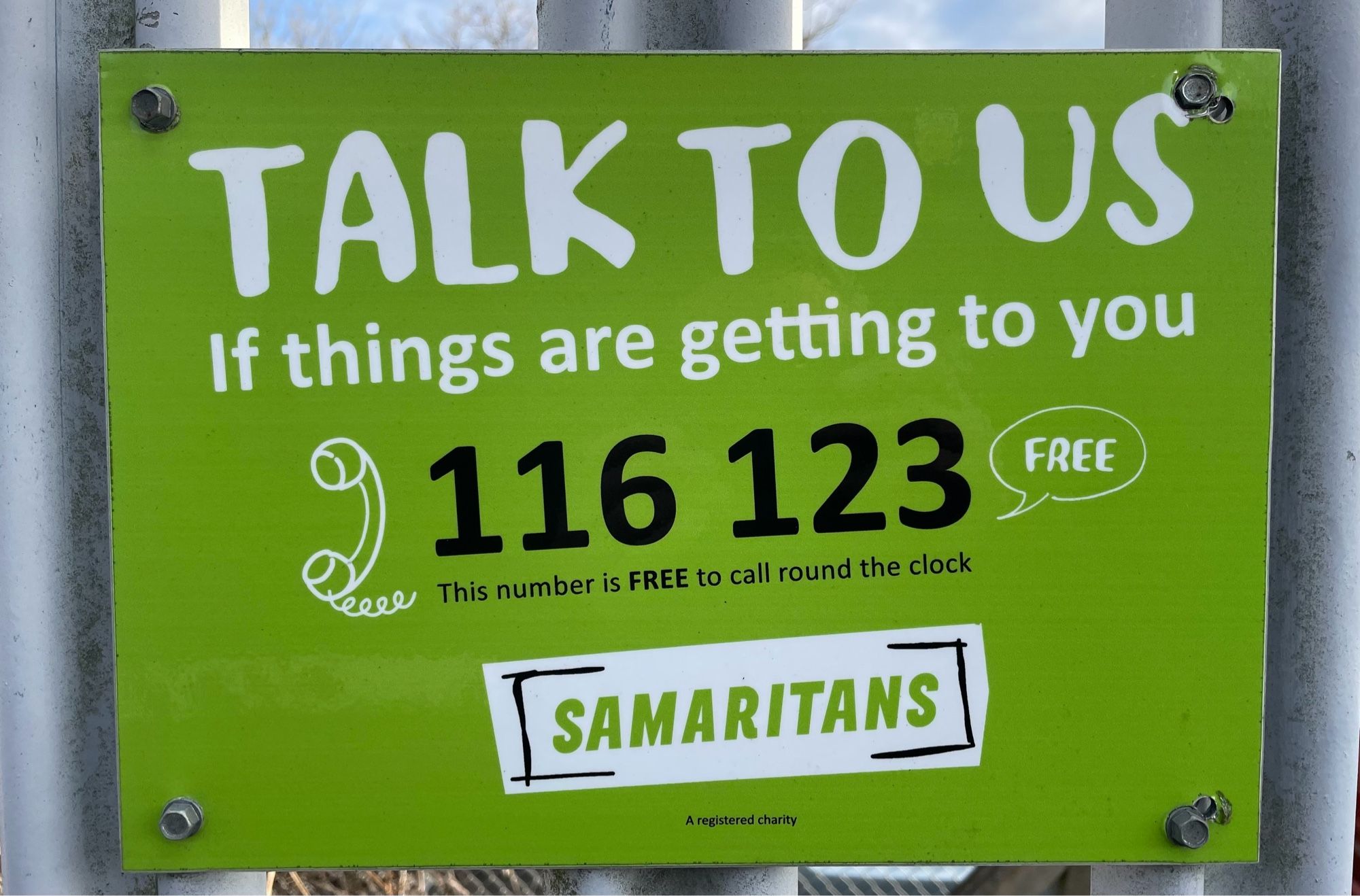 A sign affixed to railings at a station. A green background with mainly white writing at a slight angle.  The text is

TALK TO US
If things are getting to you
116 123
This number is FREE to call round the clock
SAMARITANS
A registered charity