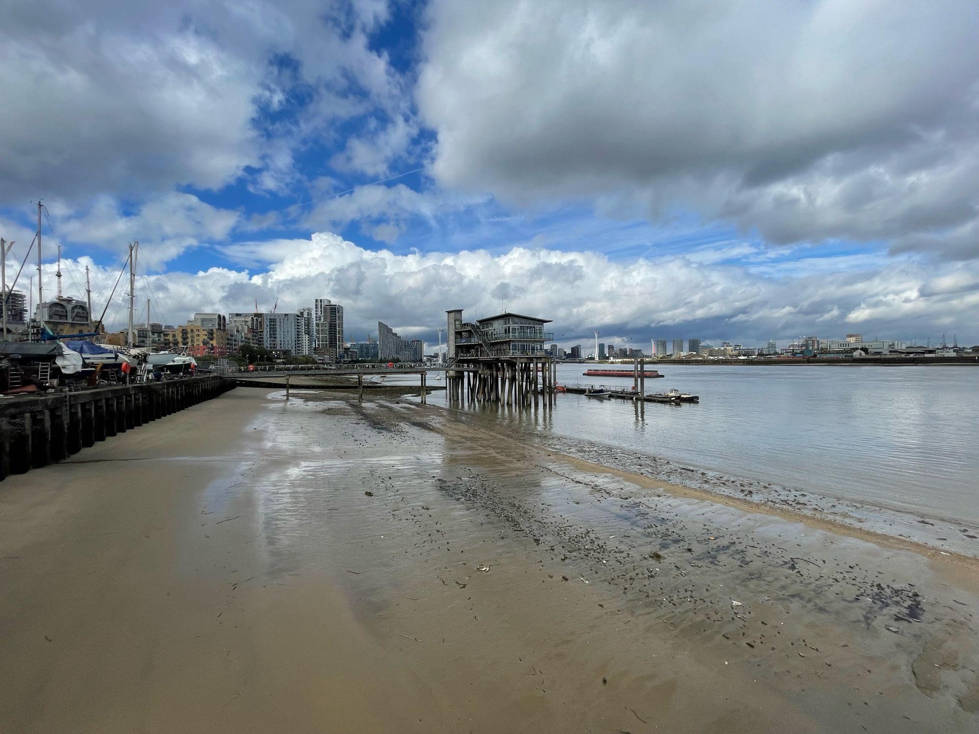 Wet sand in the foreground, with water to the right - the Thames. In the mid distance a structure on stilts - Greenwich Yacht Club, some boats from it along the left of the shot.  Towards the back medium rise blocks of flats and a the rear the high rise of Canary Wharf with the yellow supports of the O2 arena just visible.