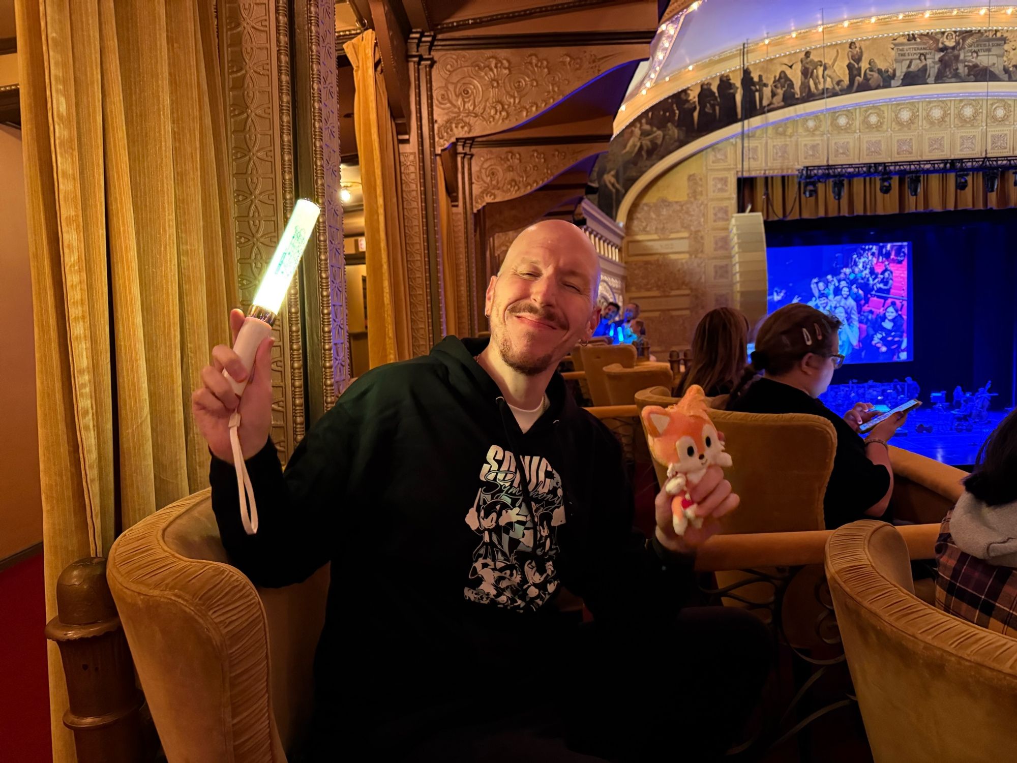 A selfie of me seated wearing a Sonic symphony hoodie at the symphony venue. I’m holding a yellow glow stick and a tails plushie.  